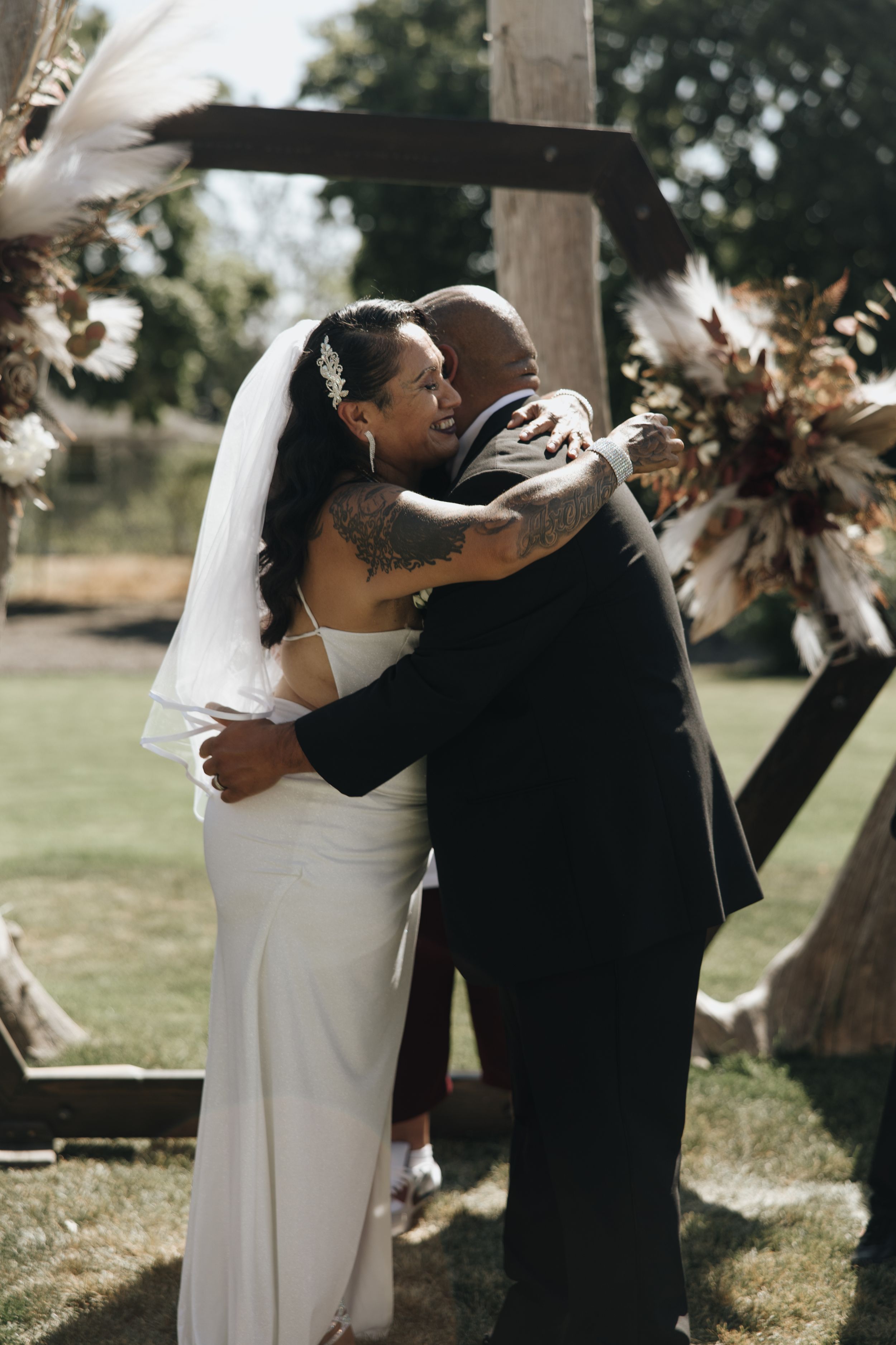 An emotional embrace during a wedding ceremony beneath a wooden arch with dried floral decorations.