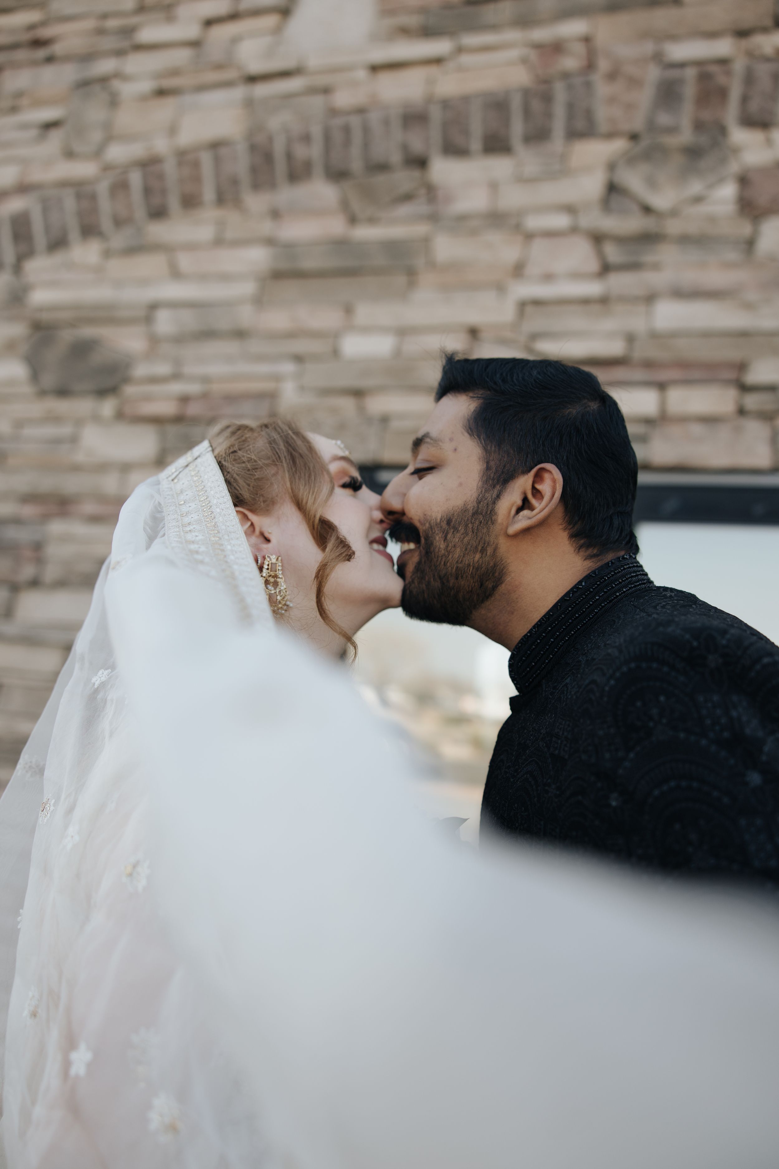 A tender wedding moment captured as a bride in her veil and groom share a romantic kiss against a stone wall.