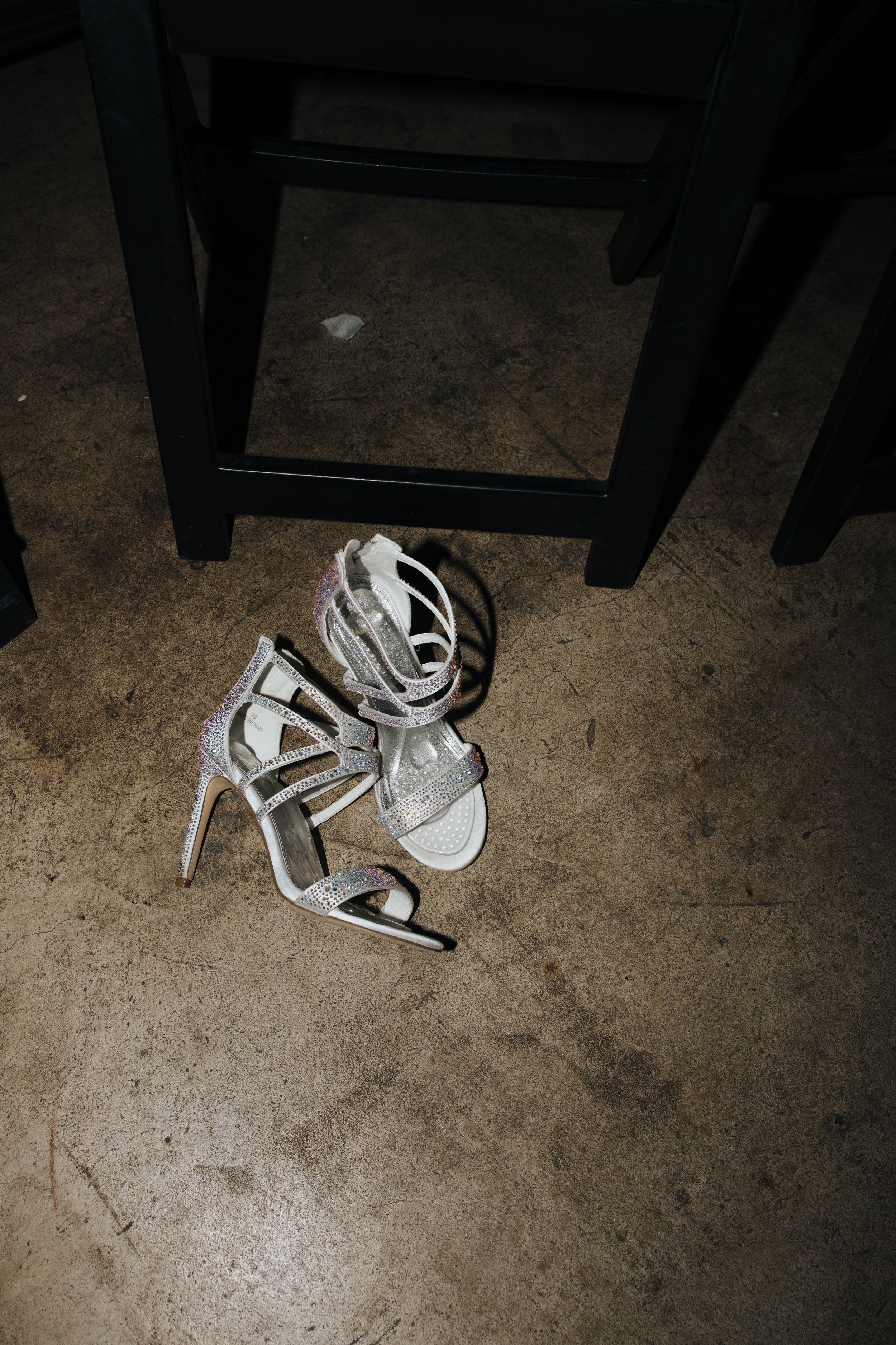 Silver strappy wedding heels discarded on concrete floor under dark furniture.