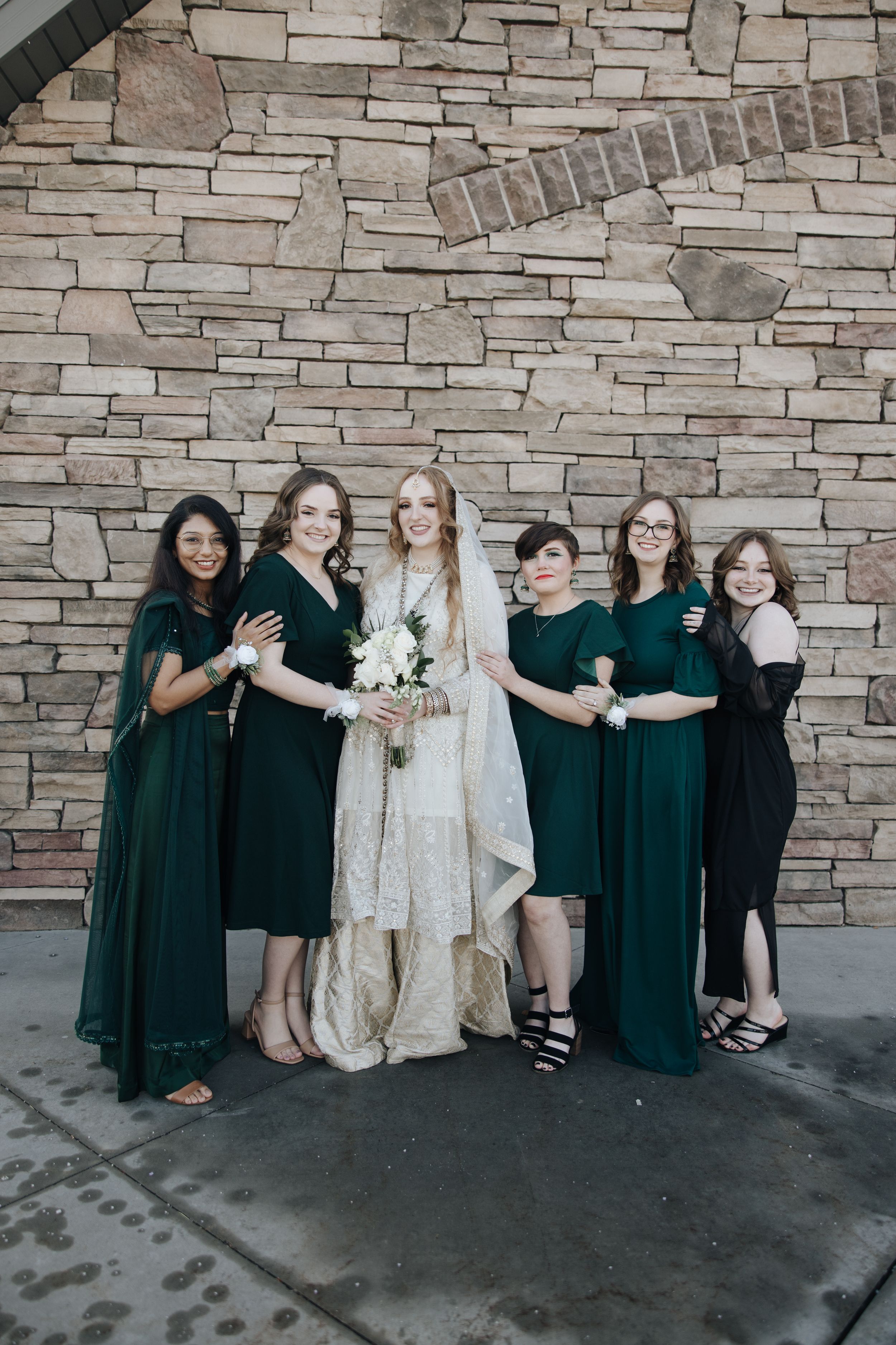 Bridal party poses against stone wall with bridesmaids wearing elegant dark green dresses.
