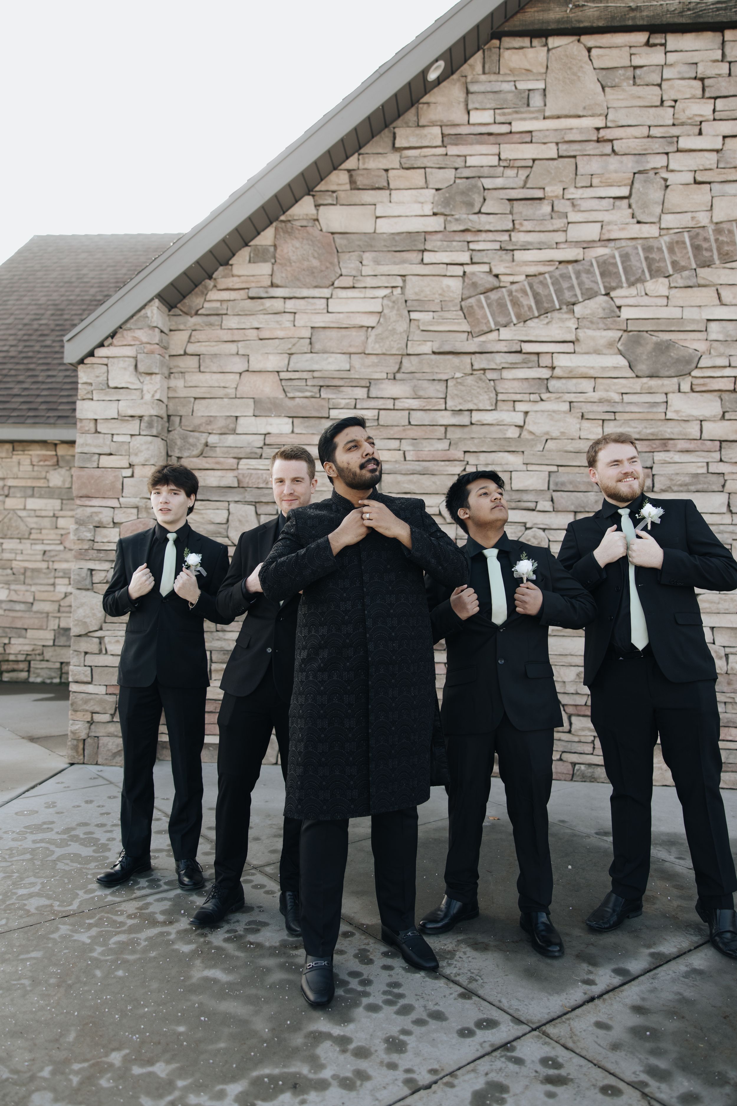 Groomsmen in black suits strike a fun pose against stone wall exterior.