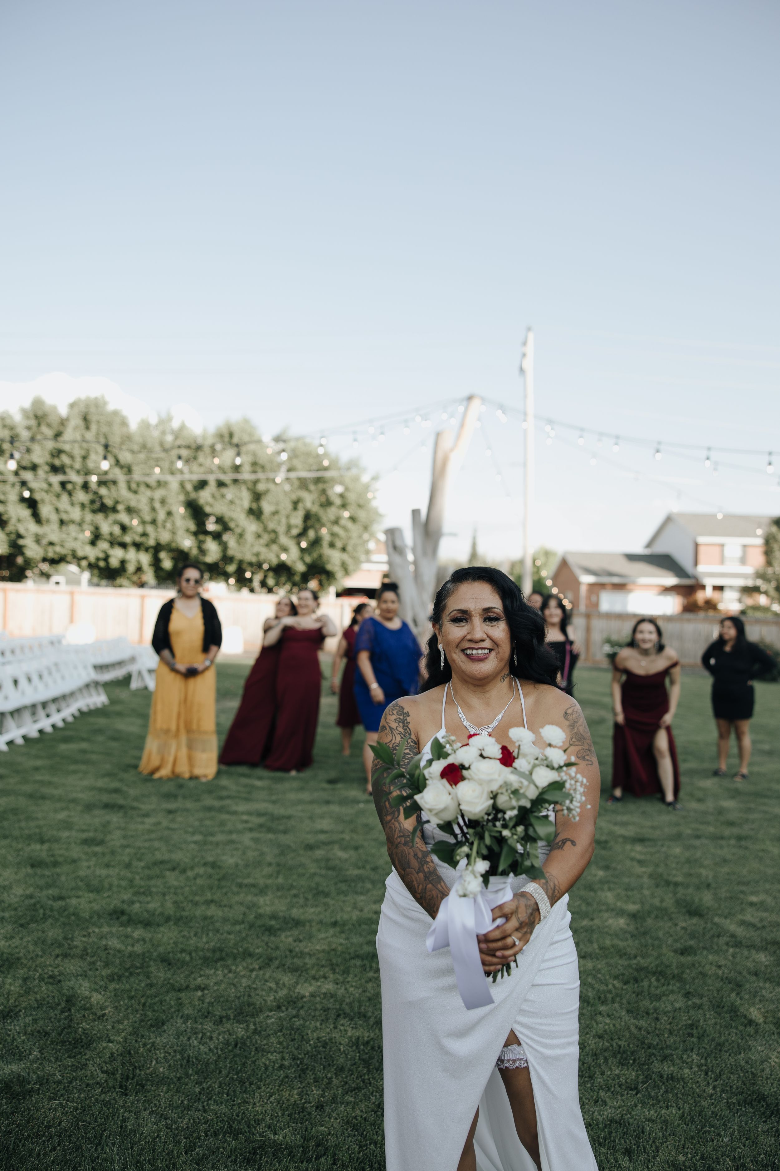 Bride in sleeveless white dress smiles while holding red and white bouquet during outdoor wedding ceremony.