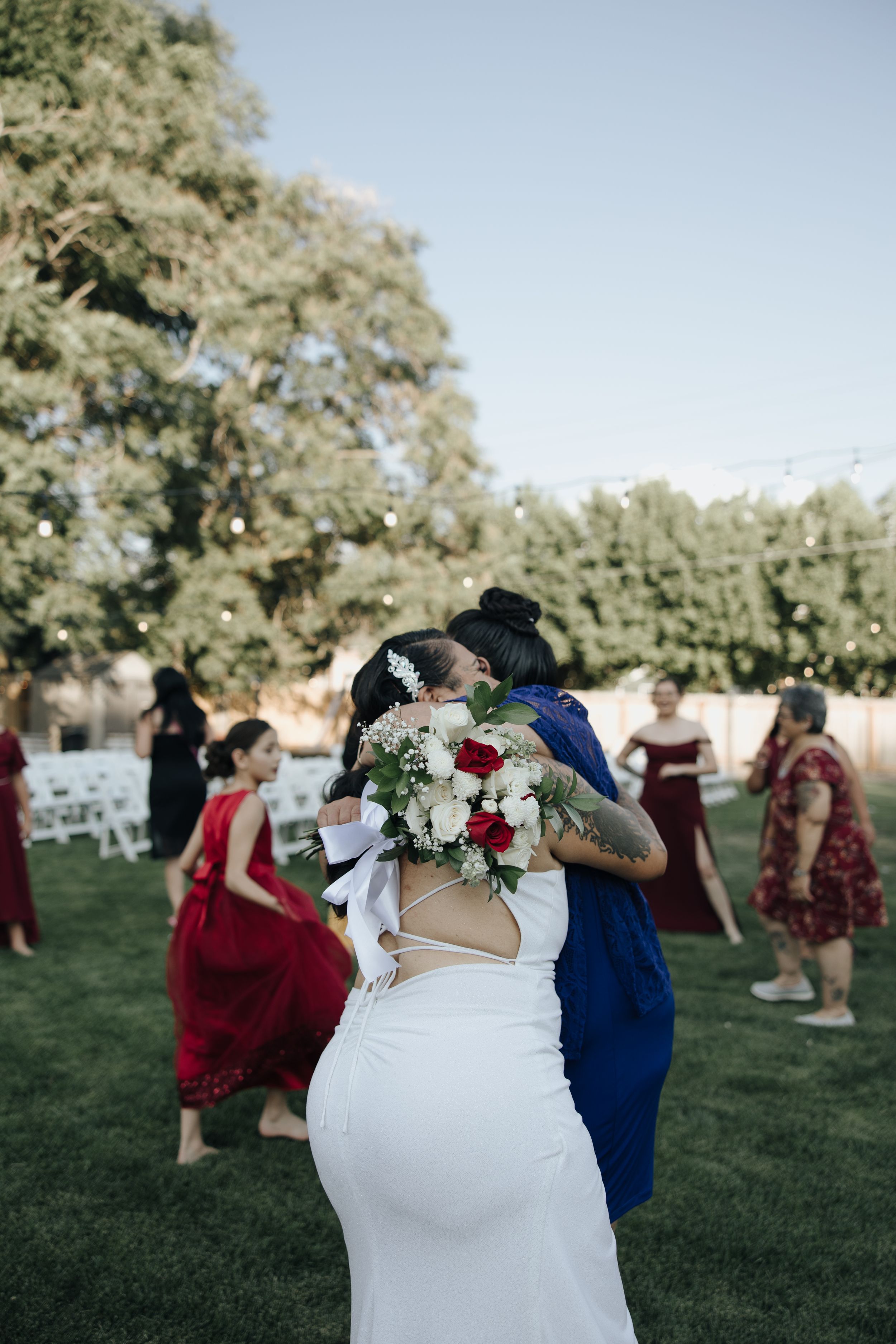 Back view of bride in white dress hugging guest during outdoor wedding reception.