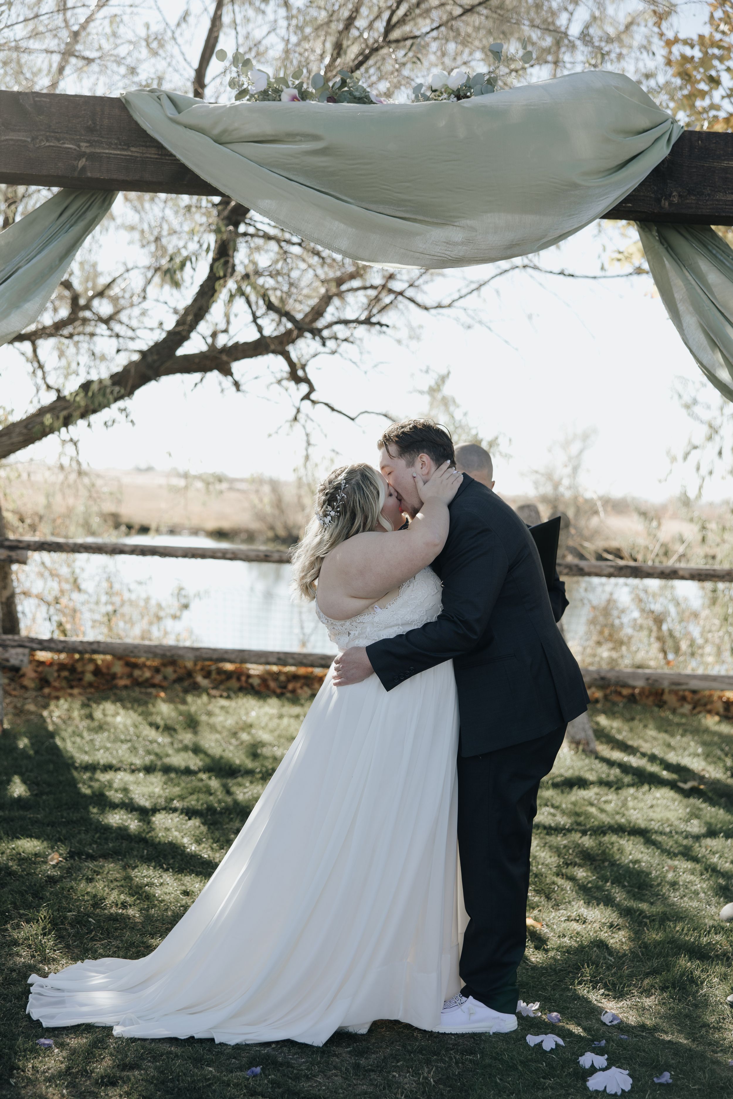 A couple shares their first kiss under a draped fabric arch at an outdoor wedding ceremony by a wooden fence.