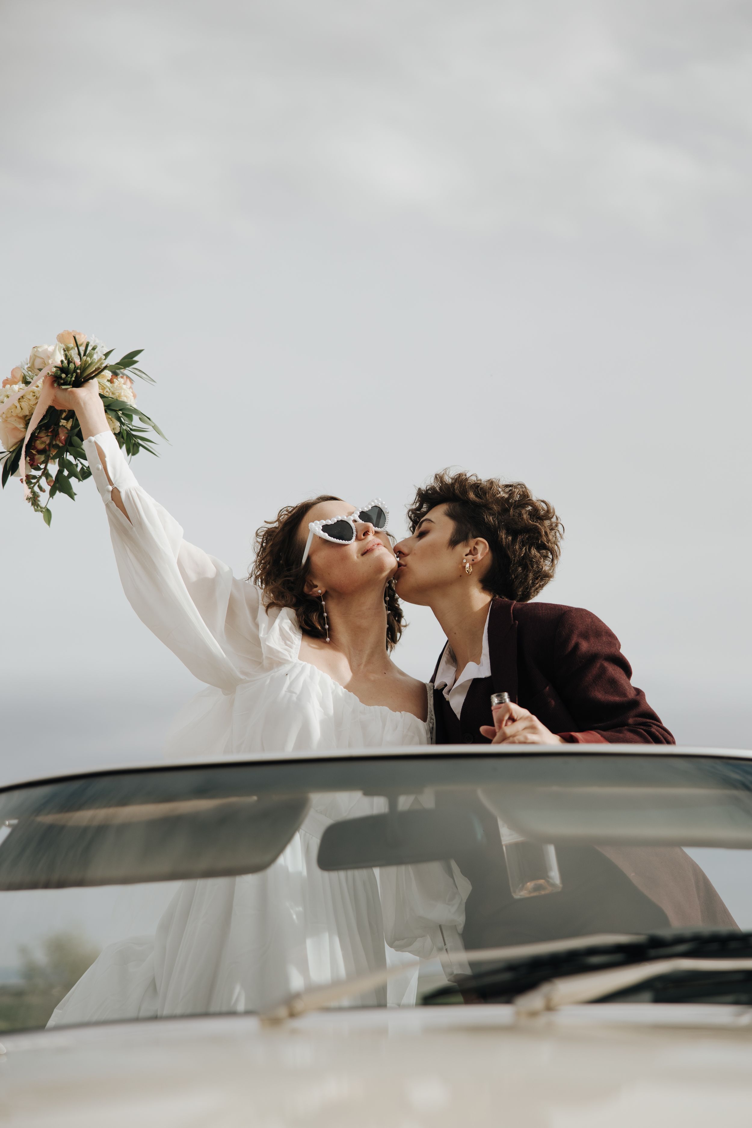 A celebratory moment through a car sunroof with a bouquet raised high against a cloudy sky.