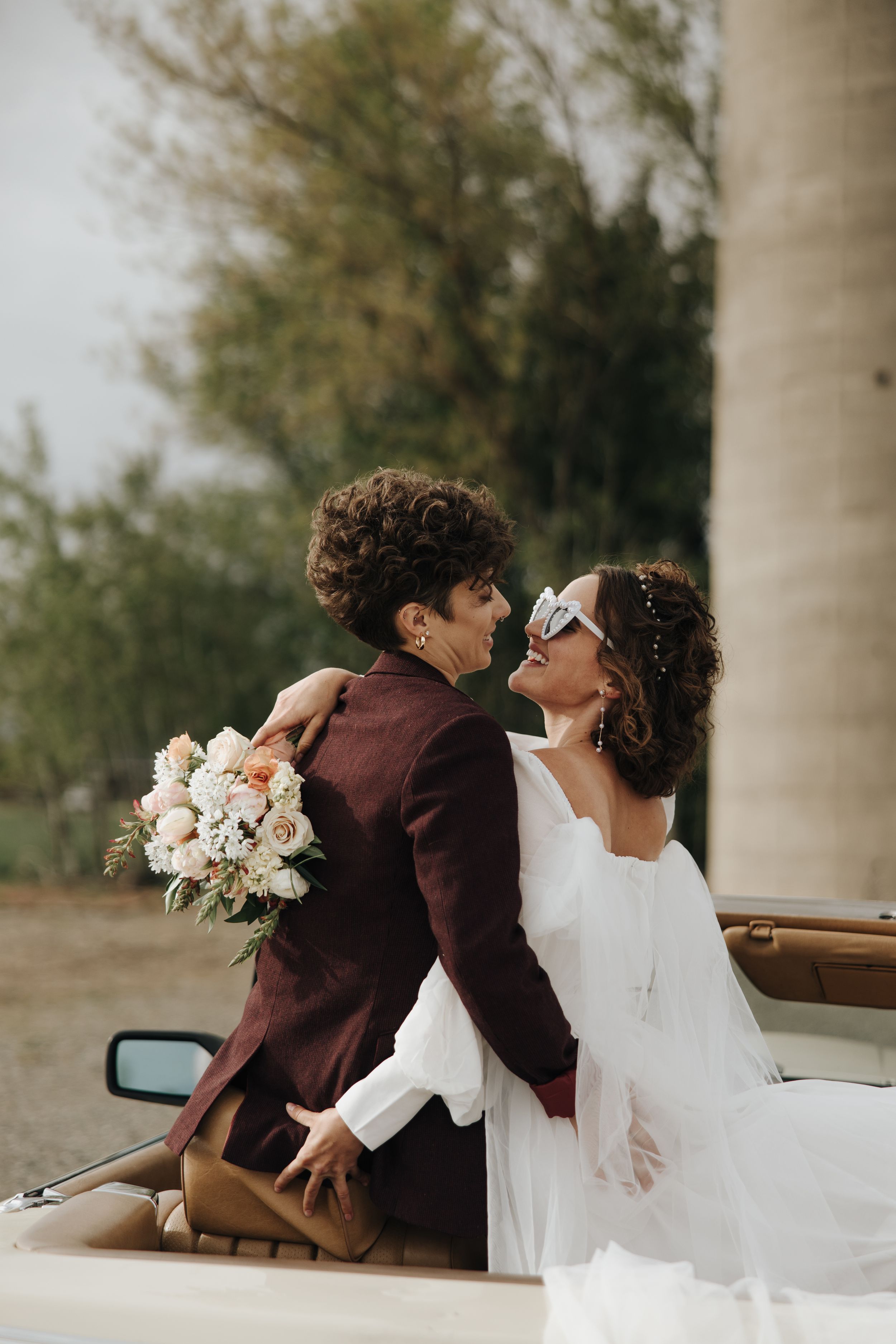 A romantic pose beside a classic car featuring a white dress and burgundy suit.