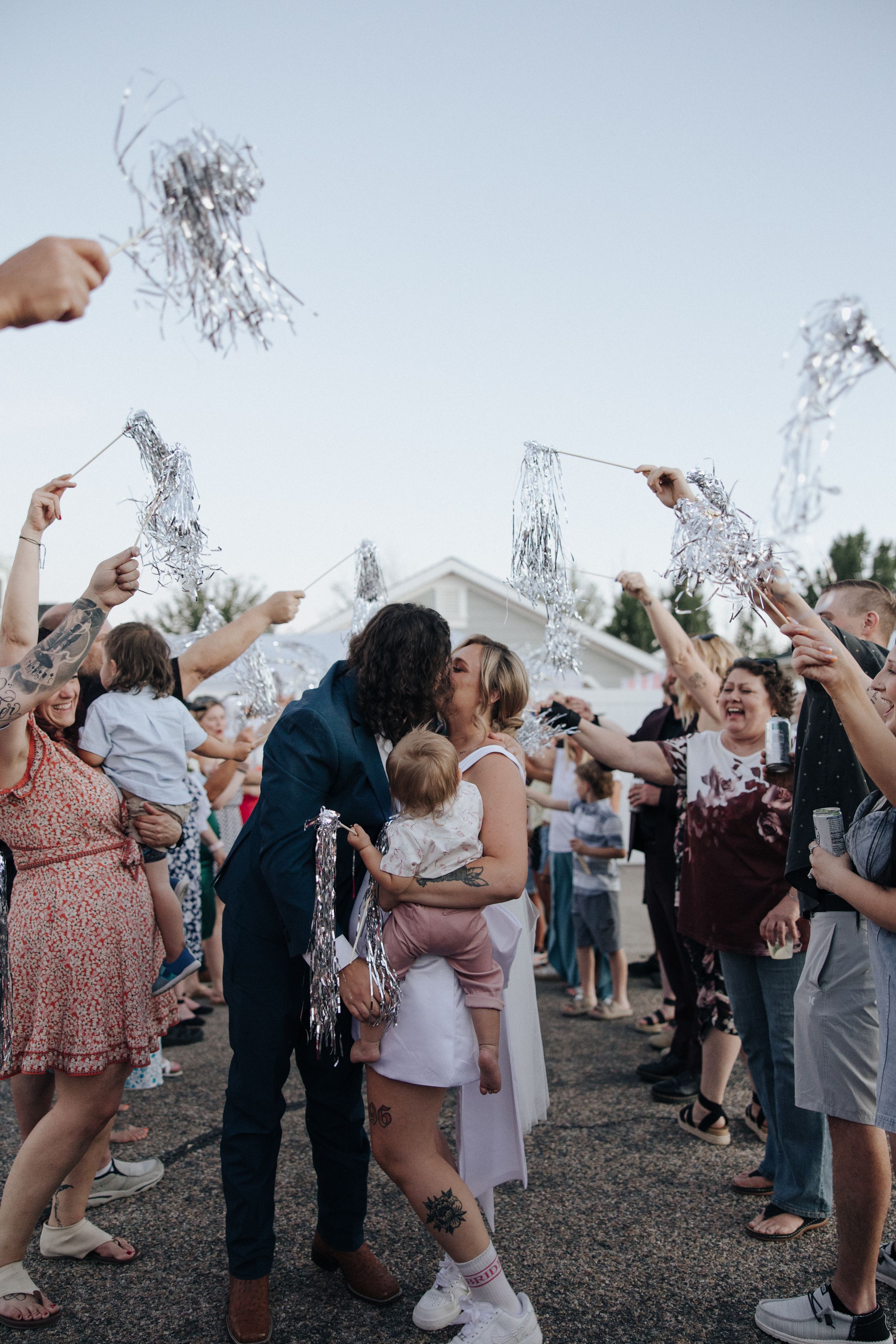 Wedding guests throw silver confetti and celebrate as couple shares romantic moment among the festivities outdoors.