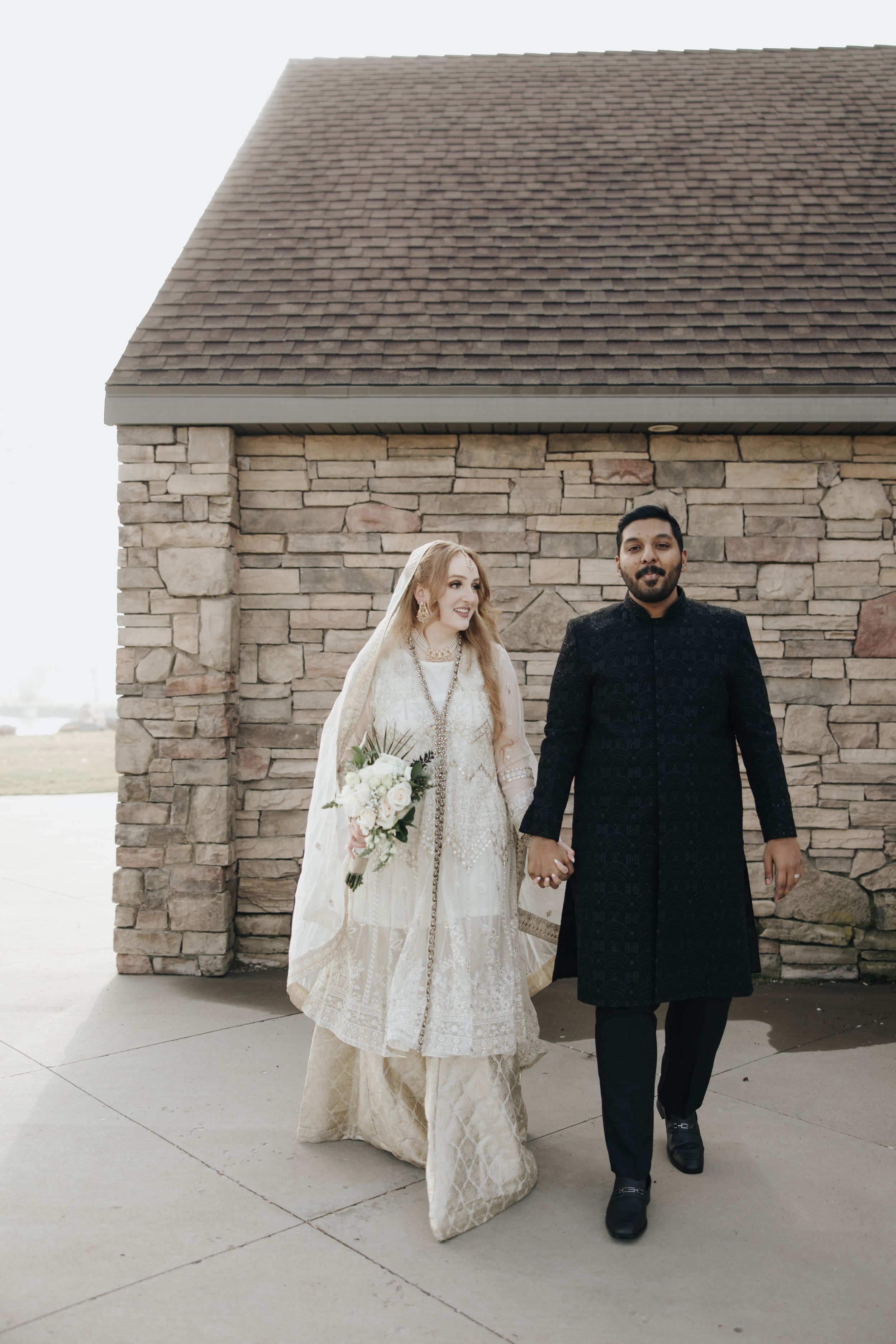 Wedding couple walks together against stone building backdrop in traditional dress.