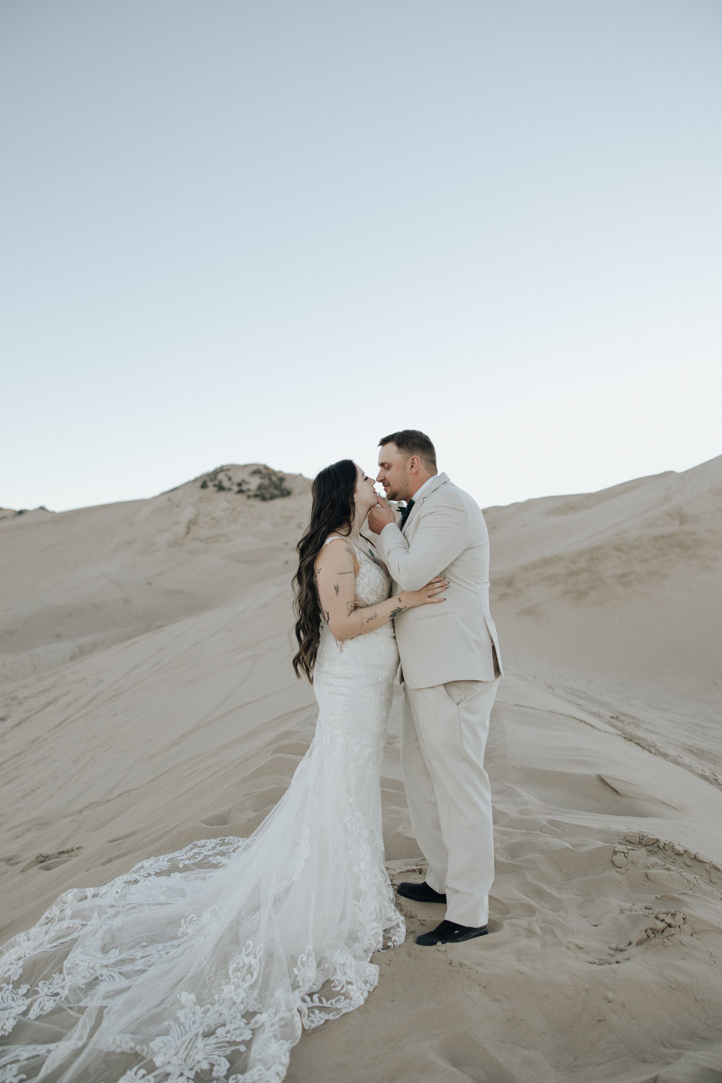 A couple in wedding attire shares a romantic moment on sandy dunes during a desert photoshoot at sunset.