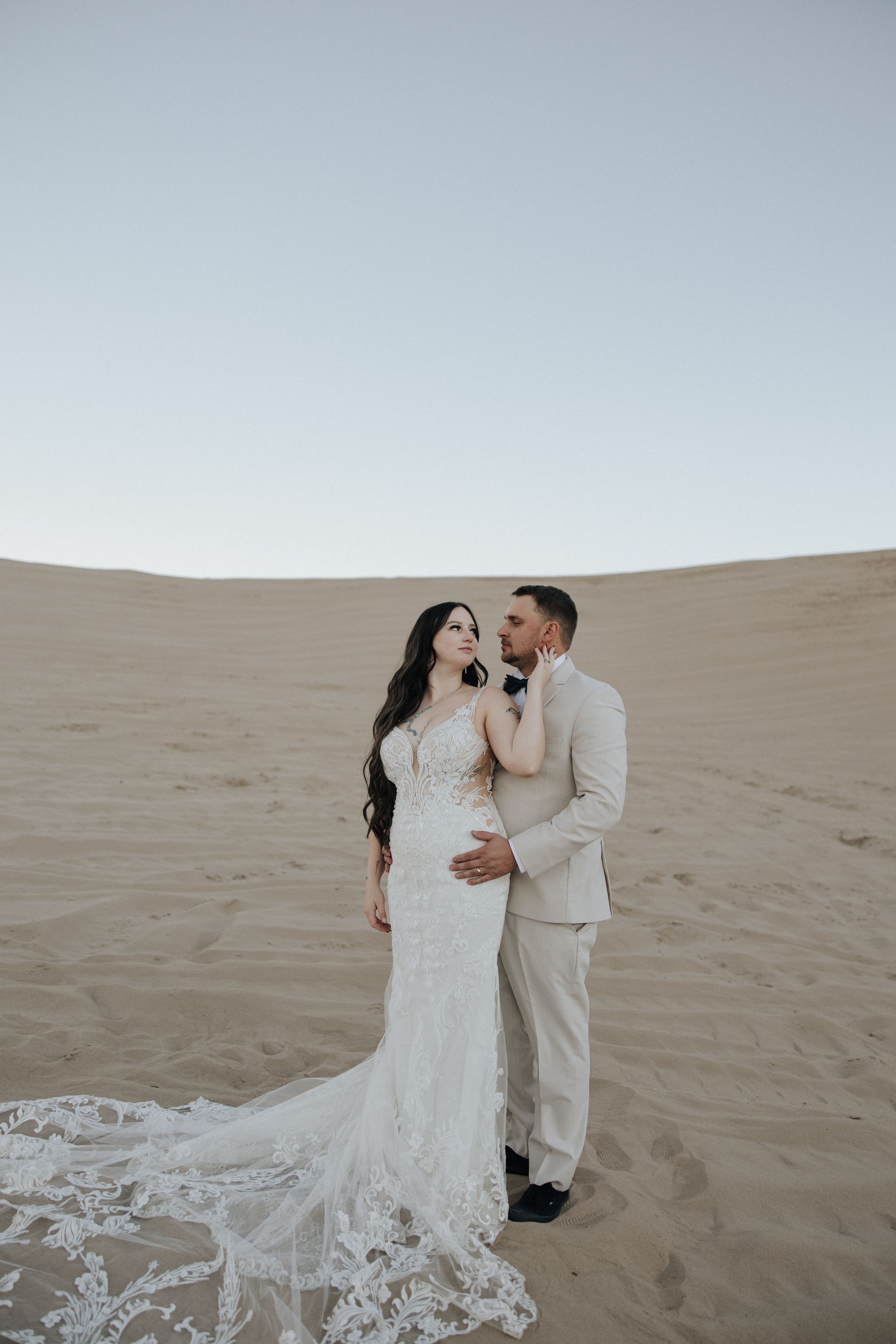 A couple shares a romantic moment in white attire against the backdrop of sweeping sand dunes in a desert setting.