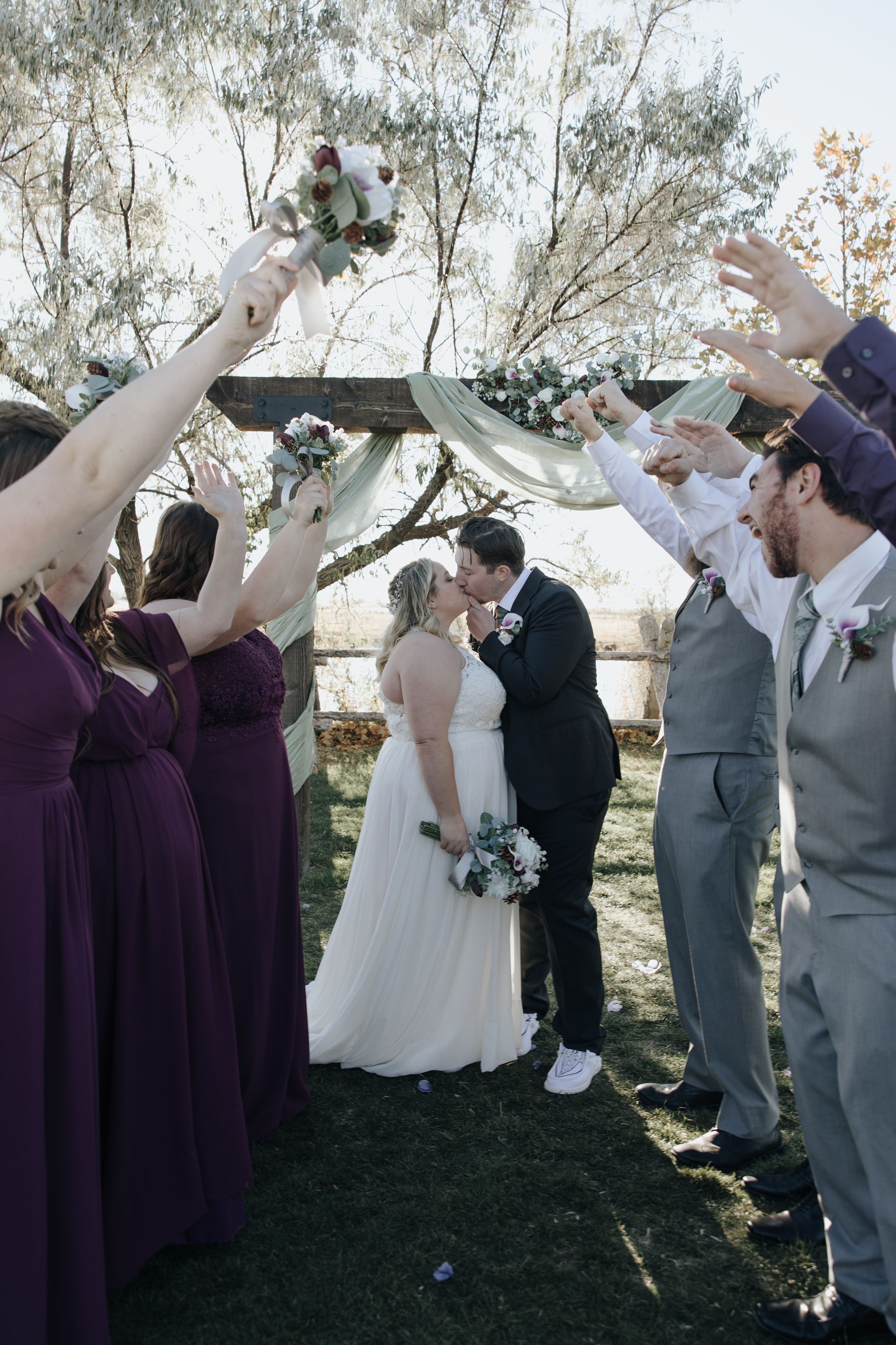 Wedding party celebrates with raised arms as newlyweds share a romantic moment under outdoor canopy.