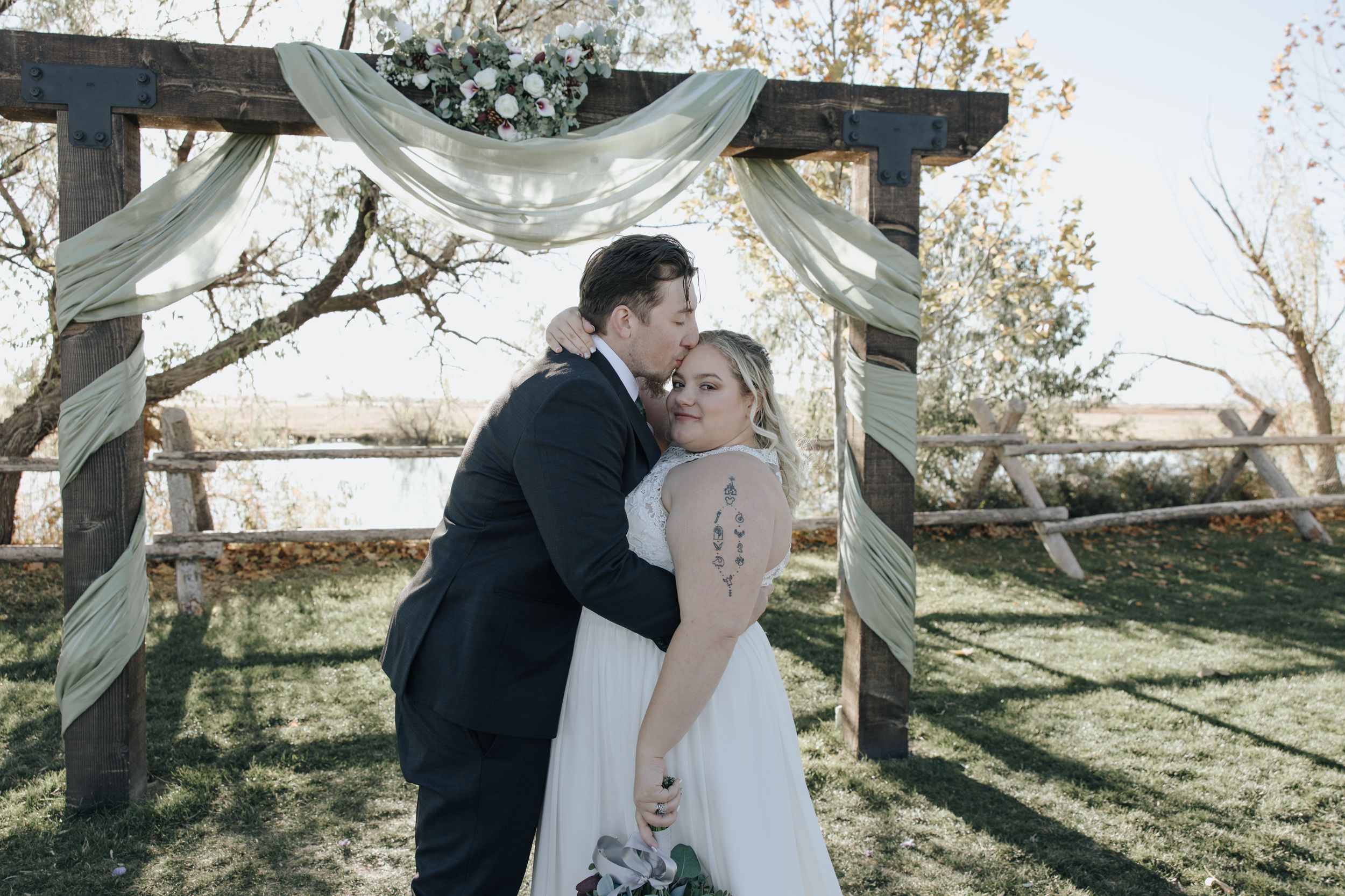 A couple sharing a tender moment under a draped arch at an outdoor wedding ceremony in autumn sunlight.