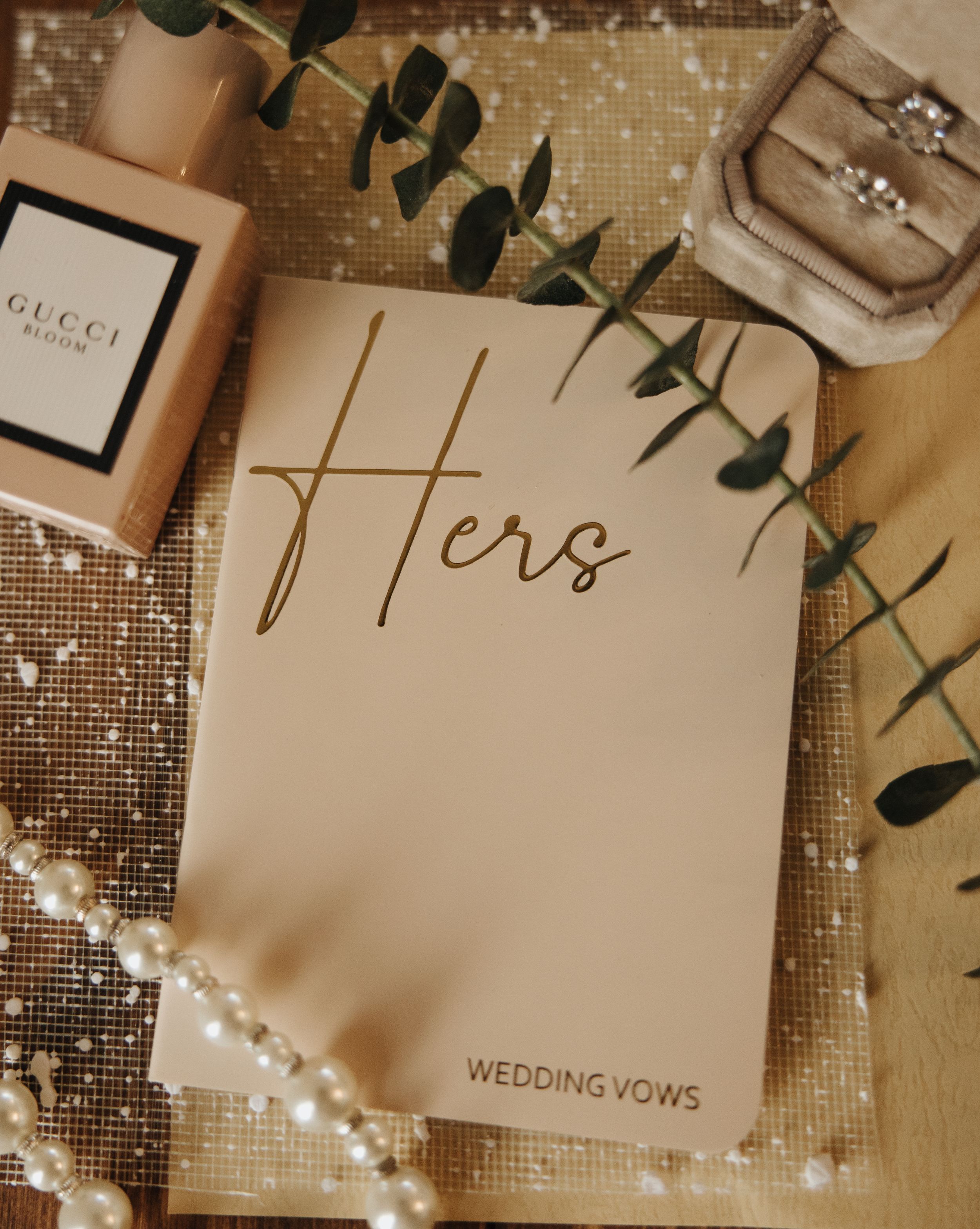 Wedding vows book with 'Hers' text alongside Gucci perfume and eucalyptus sprigs.