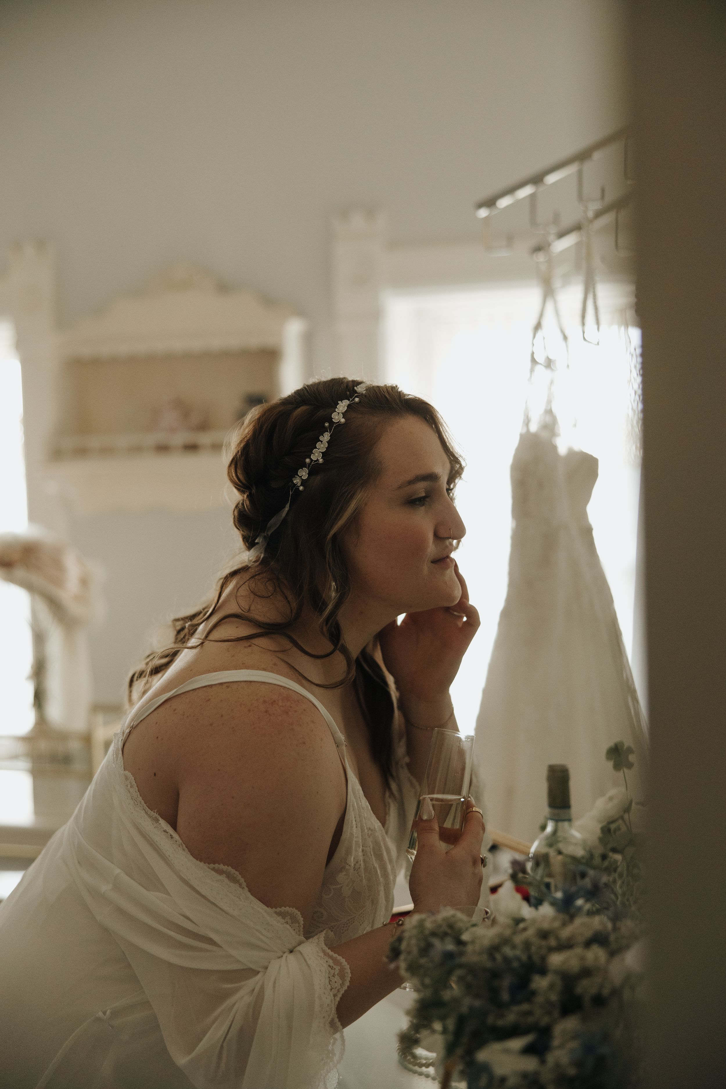 Bridal portrait in soft window light with wedding dress hanging in background.