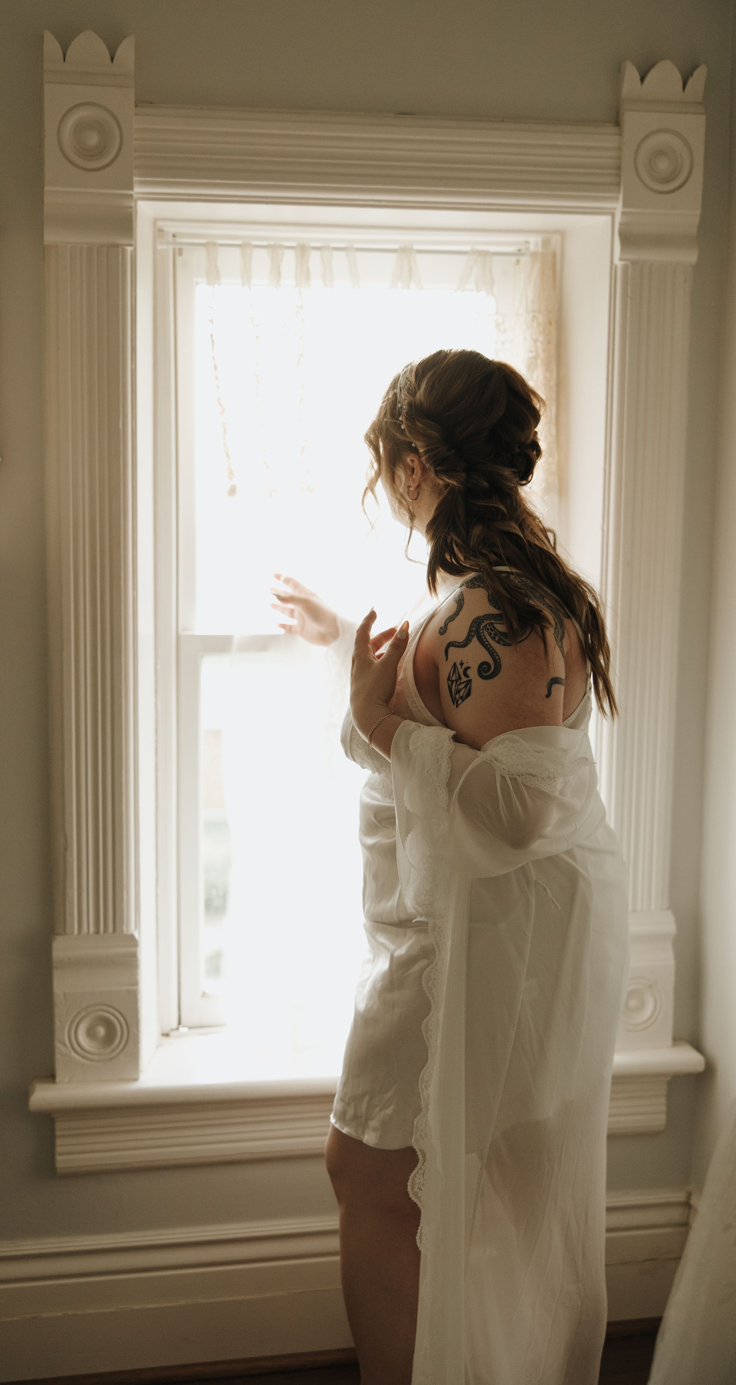 Silhouette of bride in robe looking out ornate window during wedding preparations.