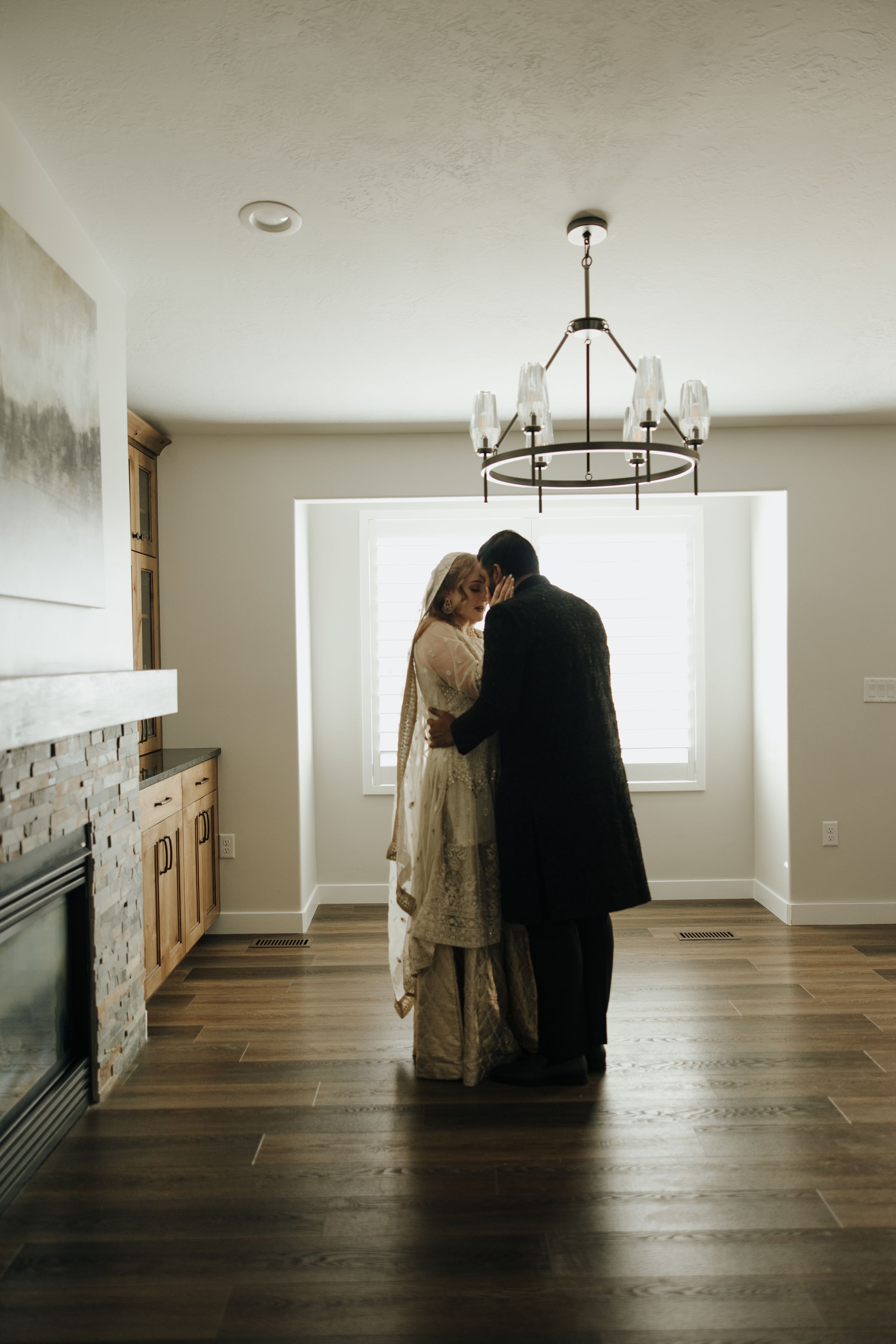 A couple embraces in silhouette against a bright window in a modern room with hardwood floors and a statement chandelier.