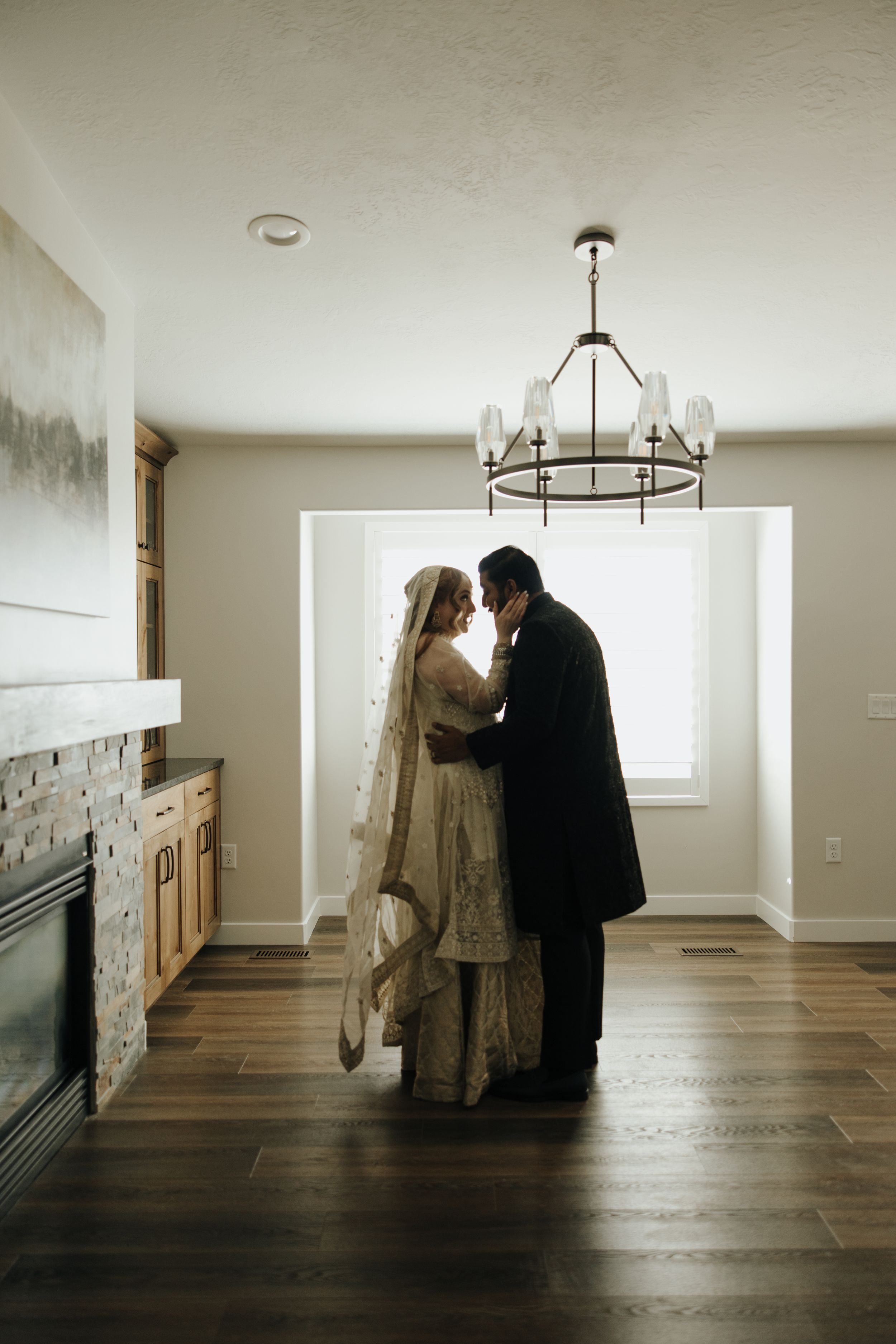 A bride and groom share an intimate moment in a modern living room with hardwood floors and chandelier lighting.