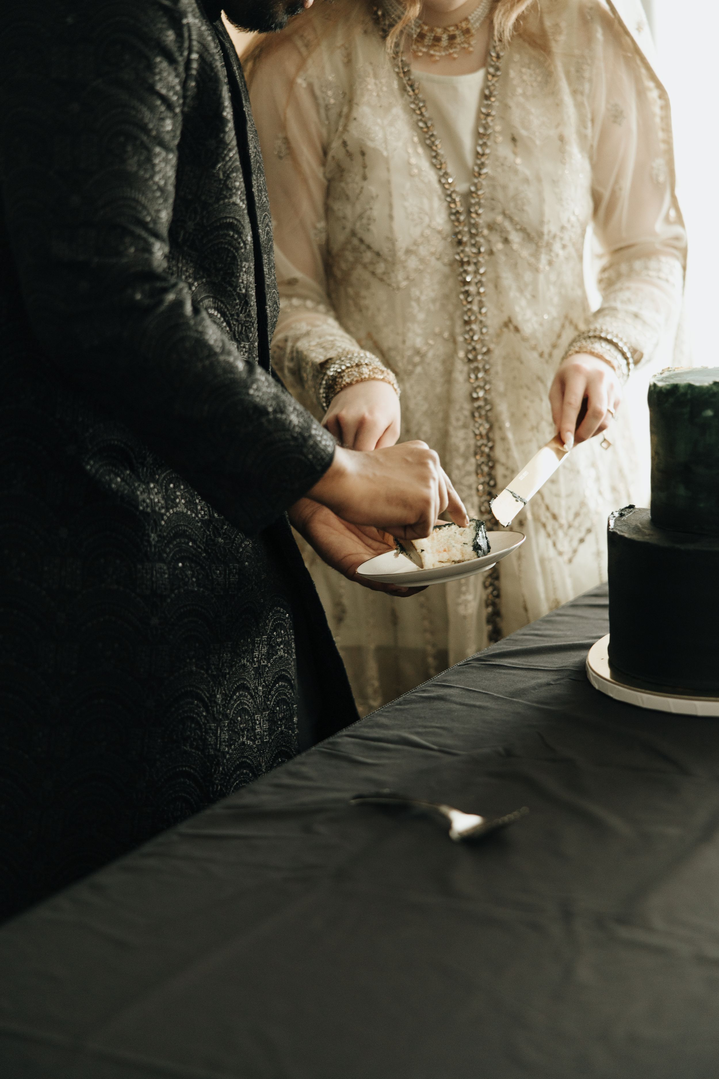 A couple in formal attire cuts into their elegant black wedding cake during an intimate celebration.