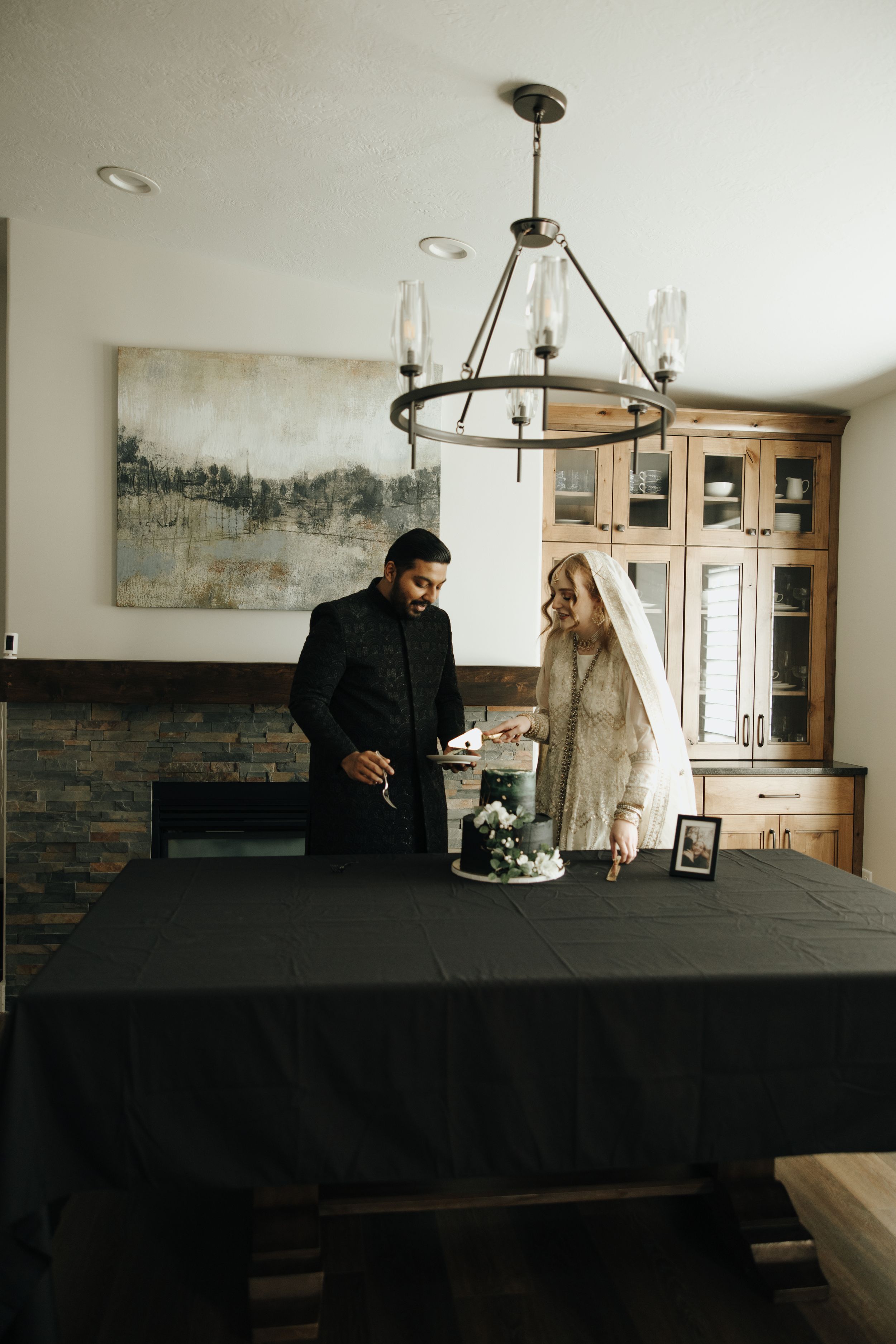 A couple stands at a dining table with a black tablecloth beneath a modern chandelier in a rustic dining room.