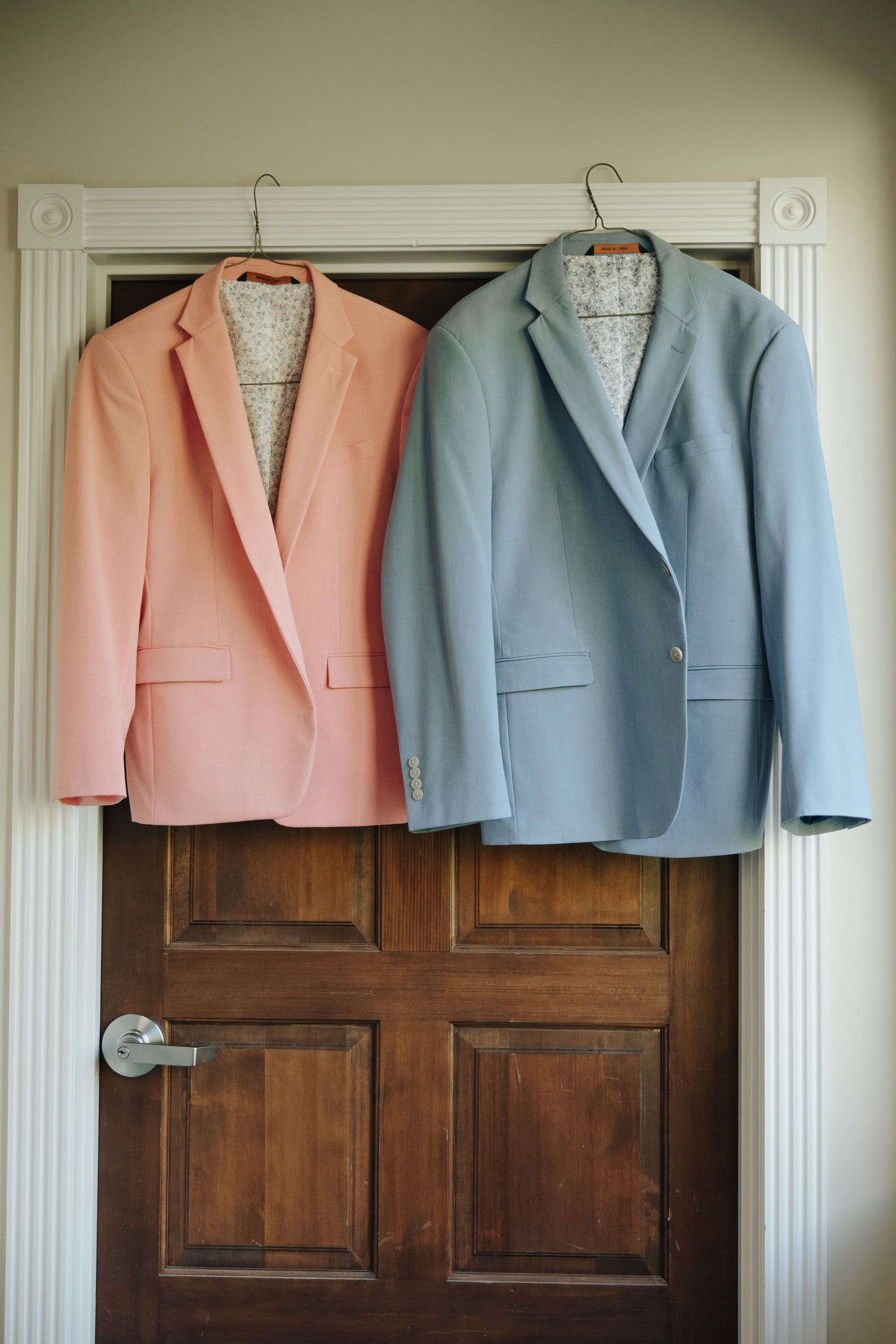 Two pastel blazers, one in peach and one in light blue, hanging on wooden hangers against a brown door.