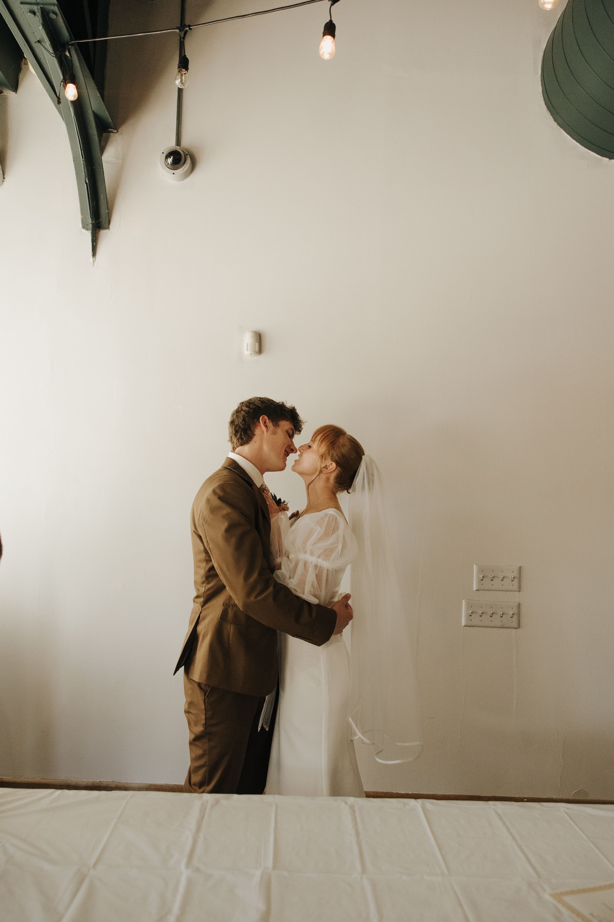 Intimate wedding moment captured against white wall with string lights.