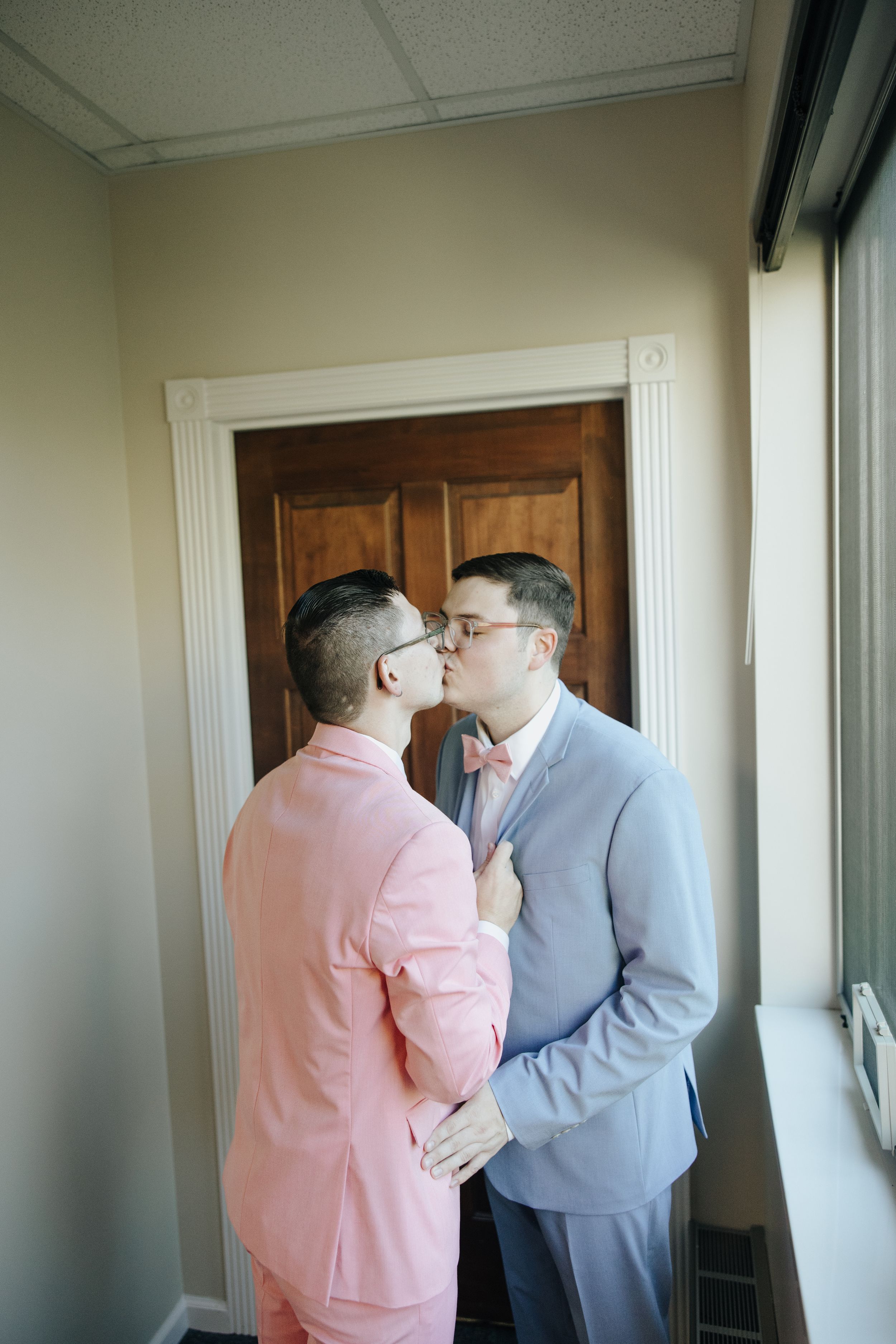 A couple wearing a pink shirt and grey suit embrace by a window and wooden door.
