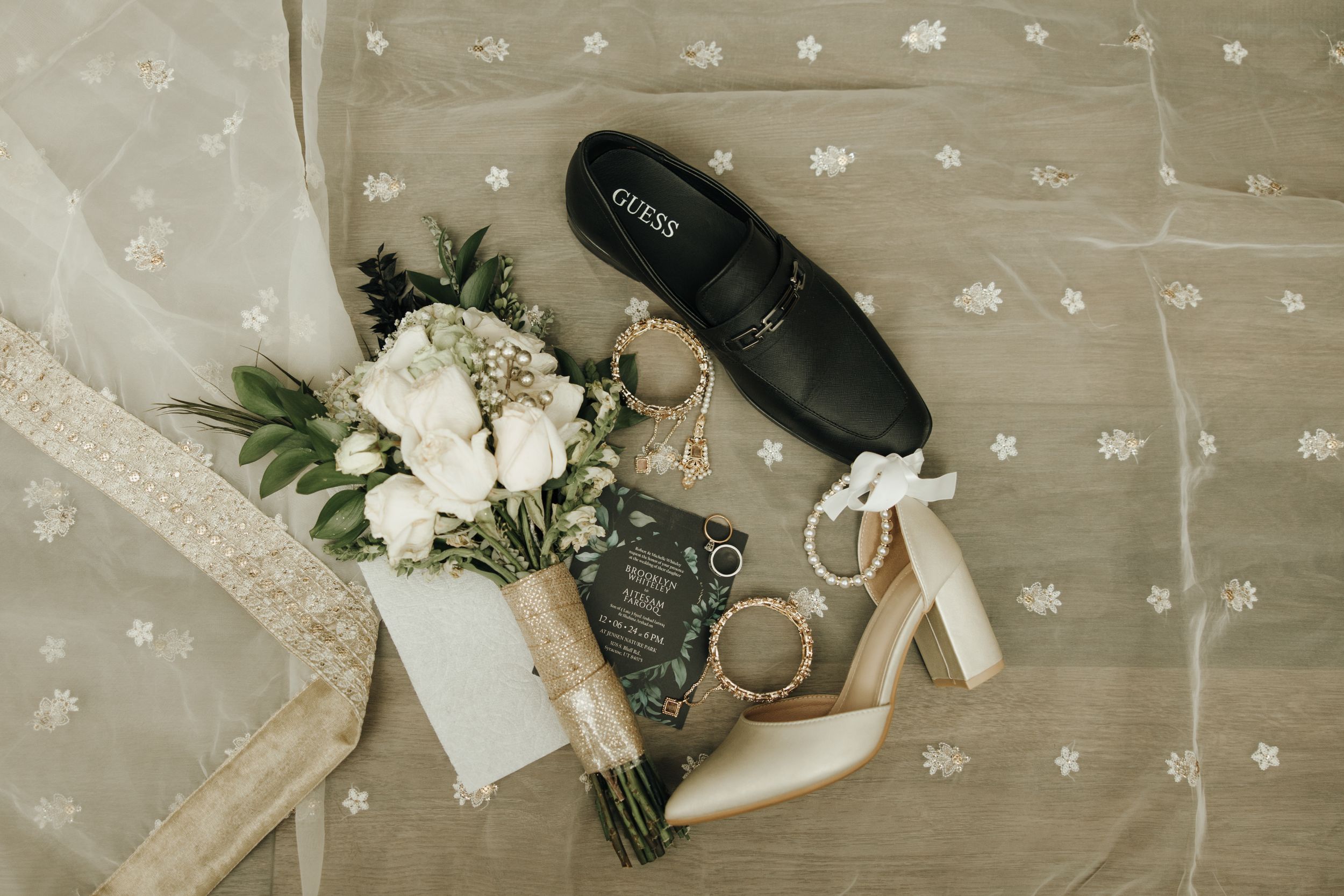 Wedding details flatlay with white floral bouquet, heels, and rings on delicate dotted fabric.