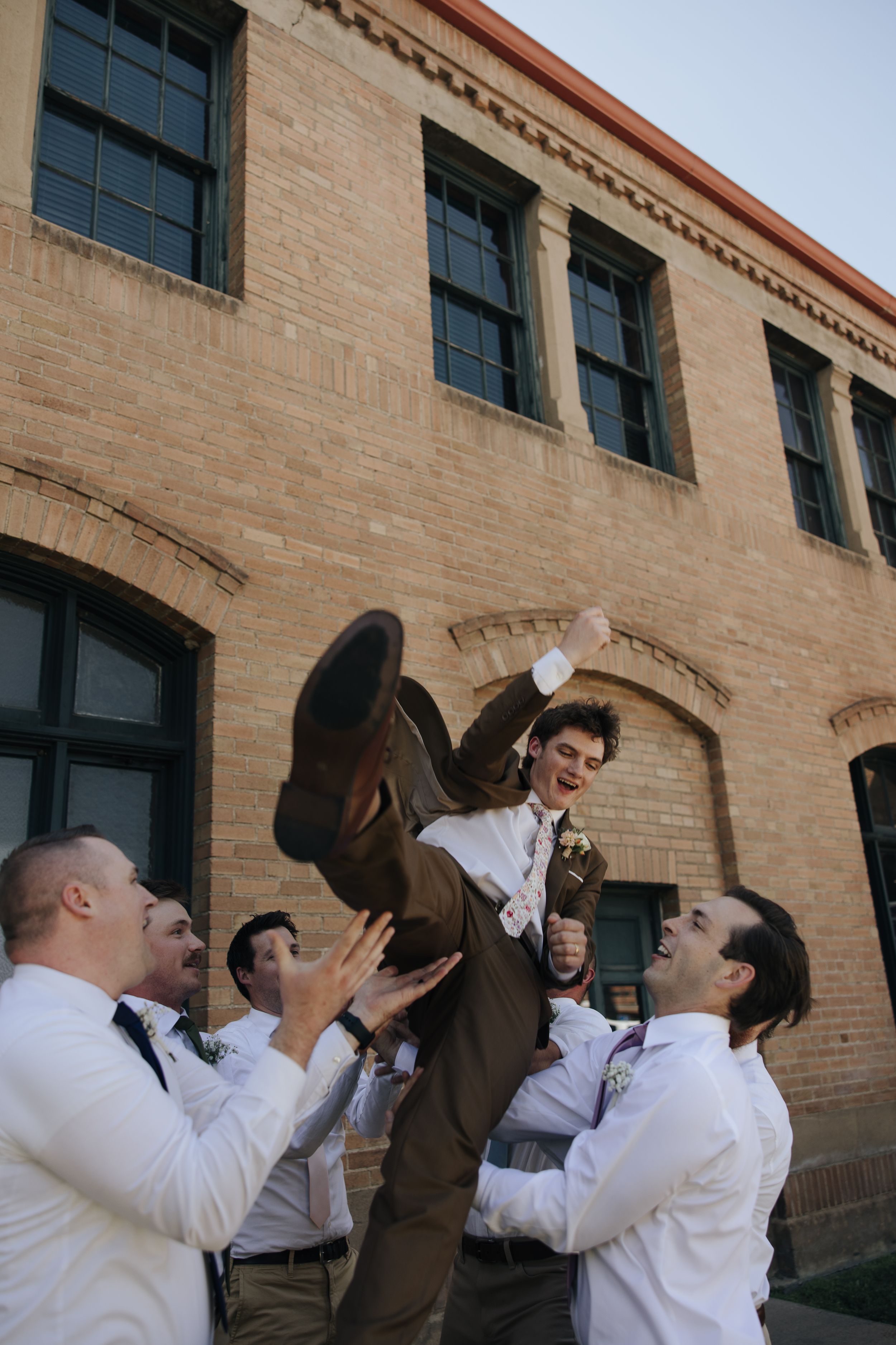 Groomsmen lifting and celebrating at a rustic brick building.