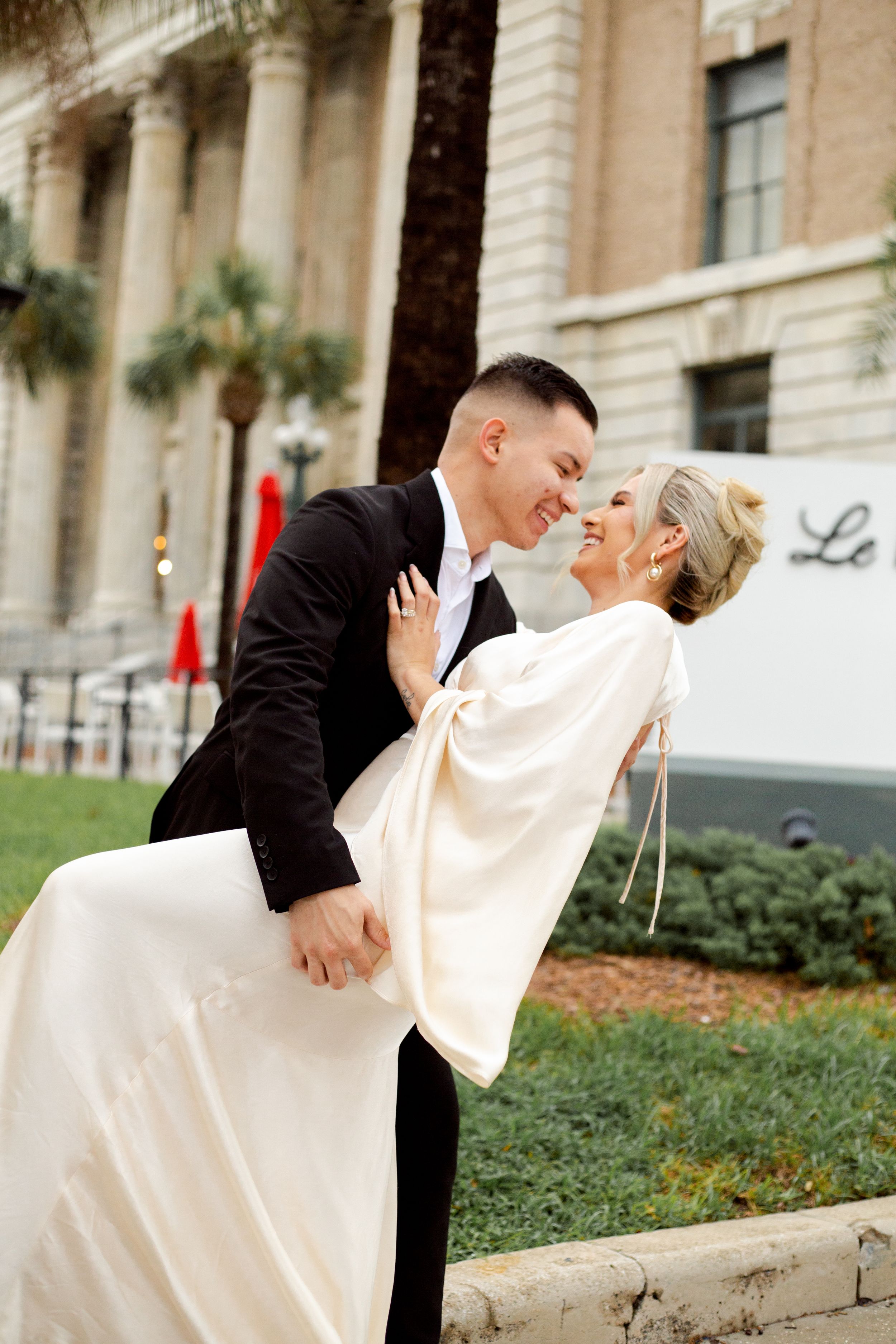 Romantic wedding couple sharing an intimate dip pose in front of a grand building with columns and palm trees.