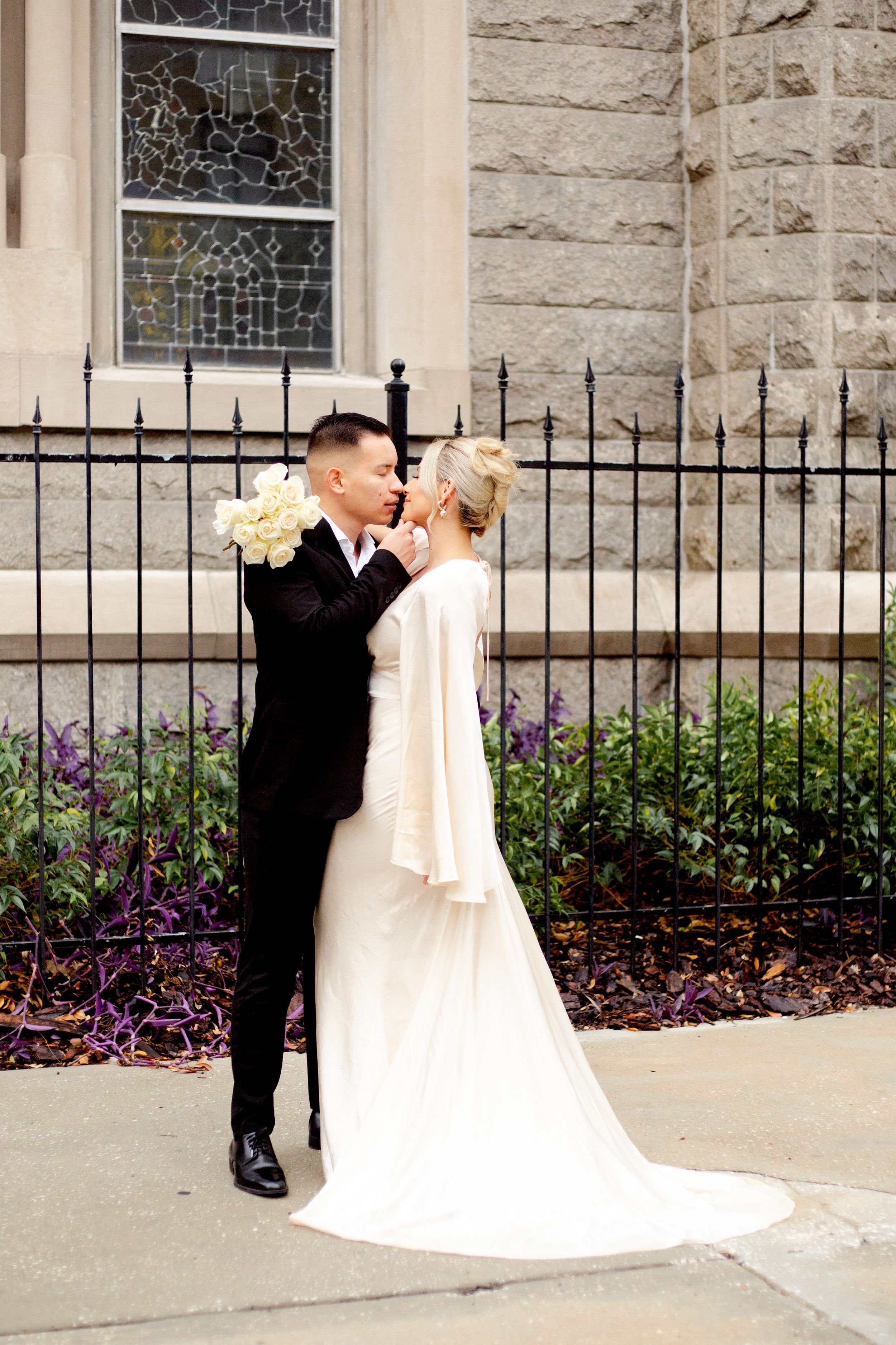 Wedding couple poses against stone building with iron fence and yellow bouquet.