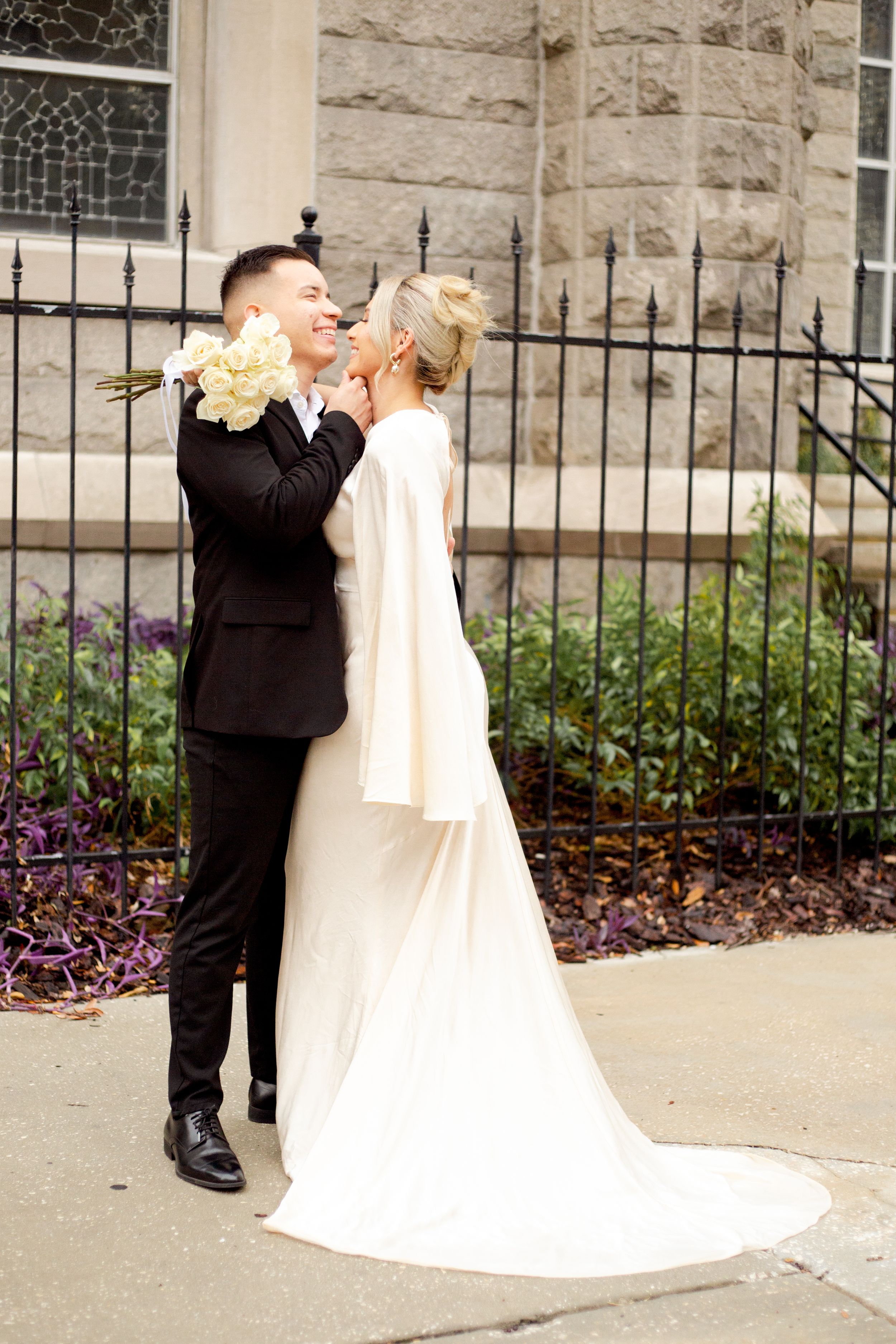 Newlywed couple embraces on church steps with yellow flowers and wrought iron fence.