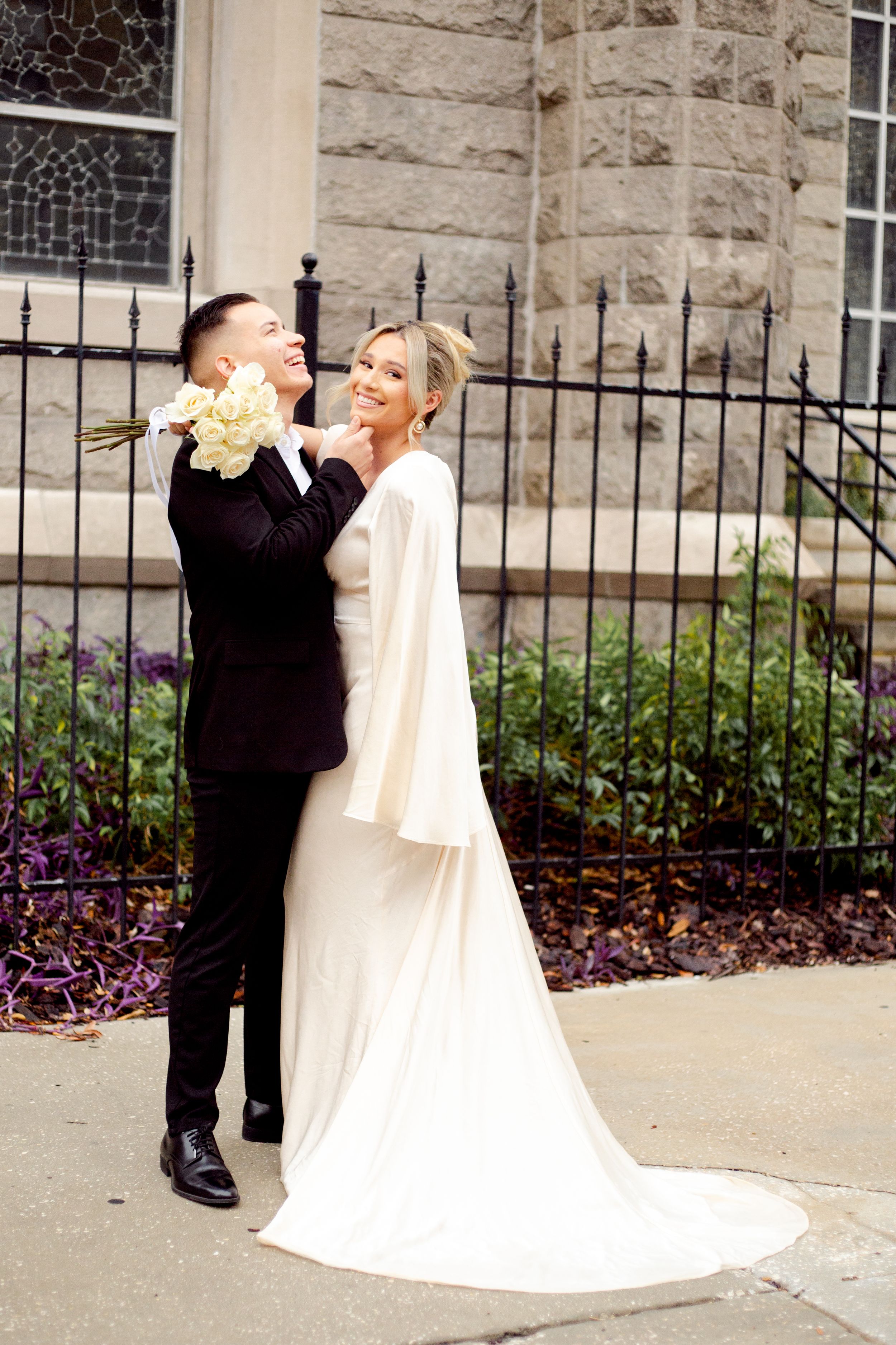 Wedding couple shares joyful moment with yellow flowers against stone church backdrop.