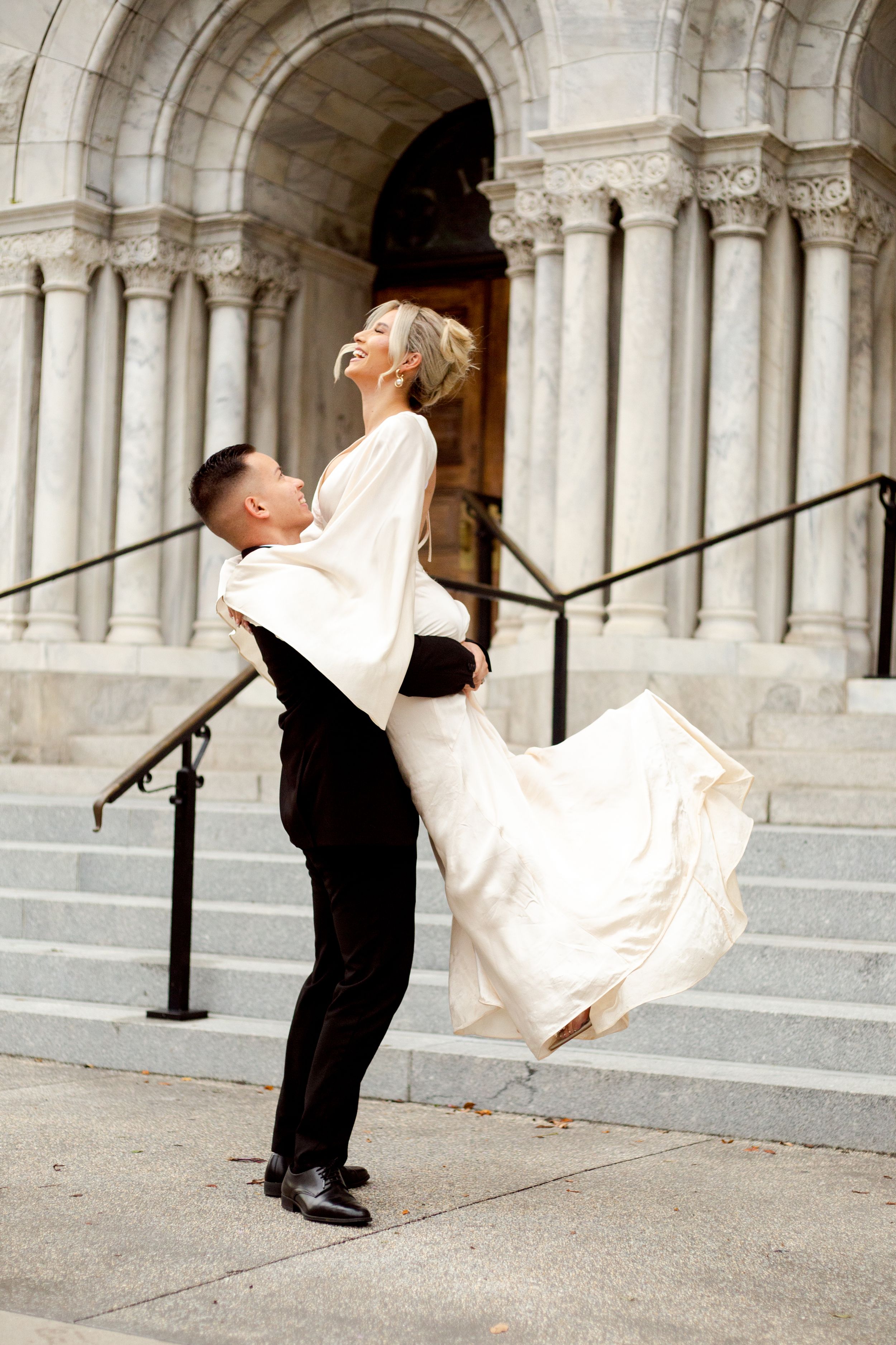 Groom lifts bride in flowing white dress on grand stone church steps.