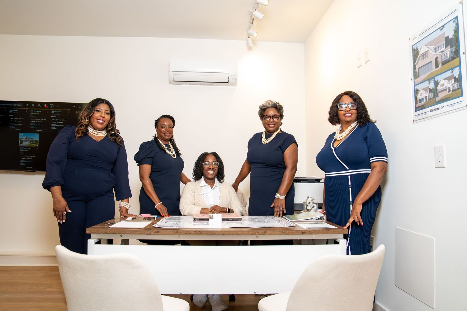 Five women in navy blue outfits gather around a desk with documents in a bright real estate office, one seated and four standing behind her.