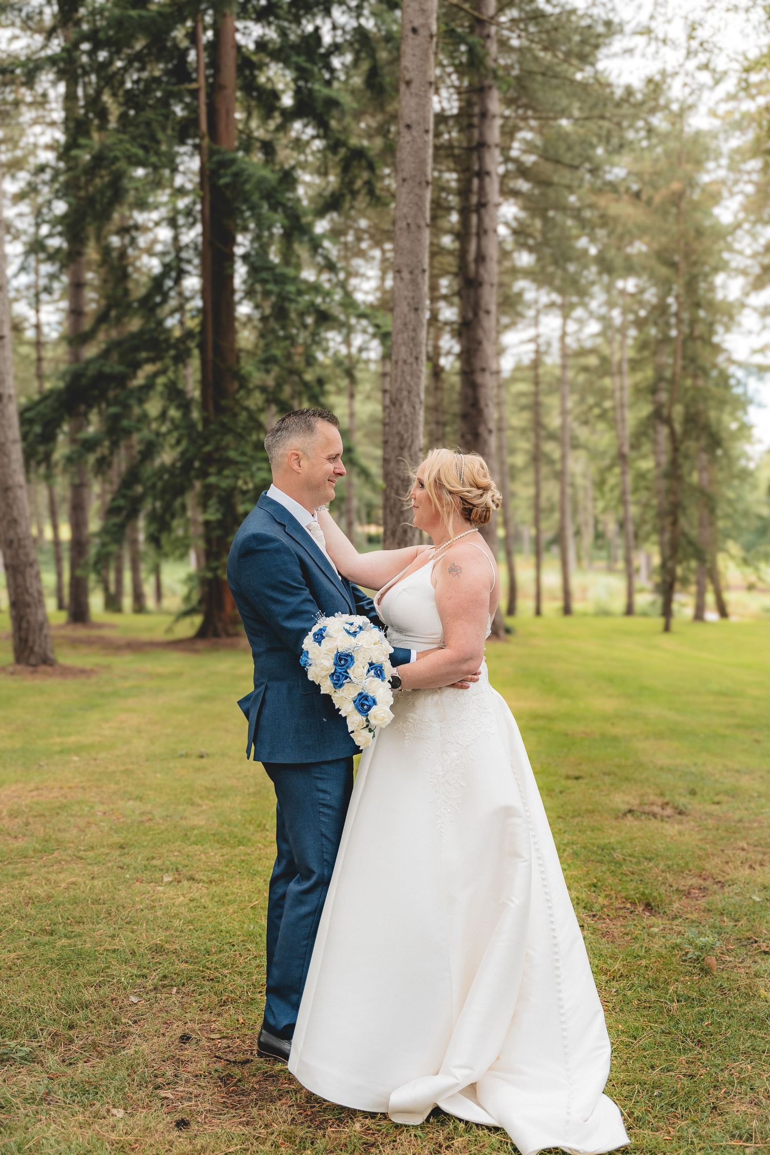 A couple in wedding attire share an intimate moment in a pine forest setting with blue floral bouquet.