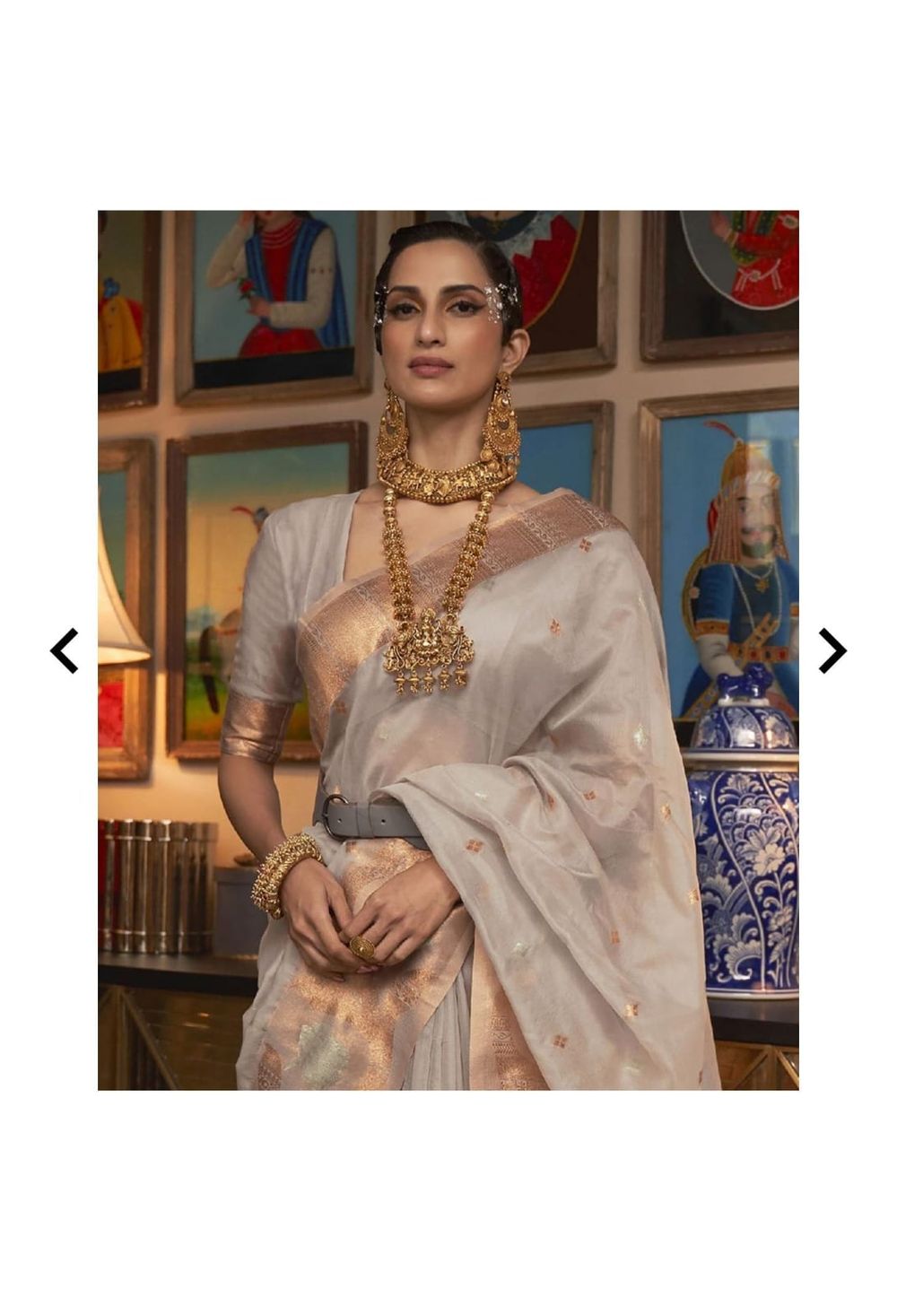 Model showcasing traditional Indian attire with heavy gold jewelry against wall art display.