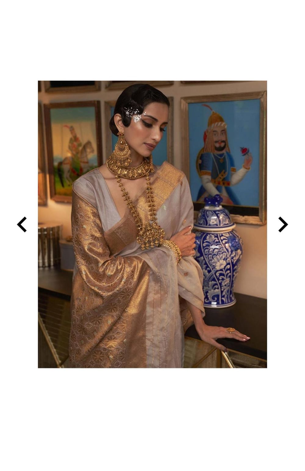 Person wearing ornate golden jewelry and cream colored silk traditional attire near decorative vases.