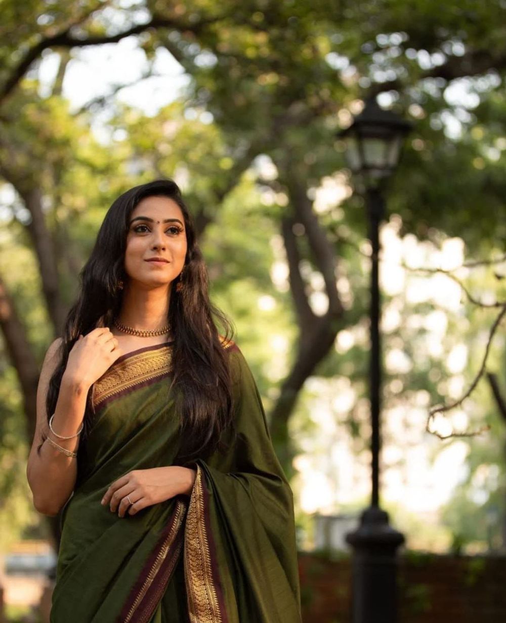 Woman in an elegant dark green saree poses gracefully in an outdoor garden setting with bokeh background.