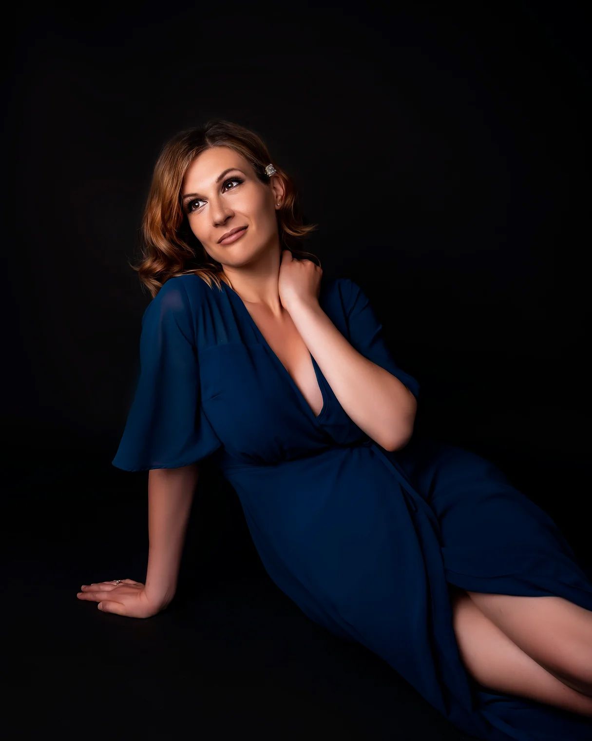 A woman in a navy blue dress poses elegantly against a dark background in a glamorous portrait session.