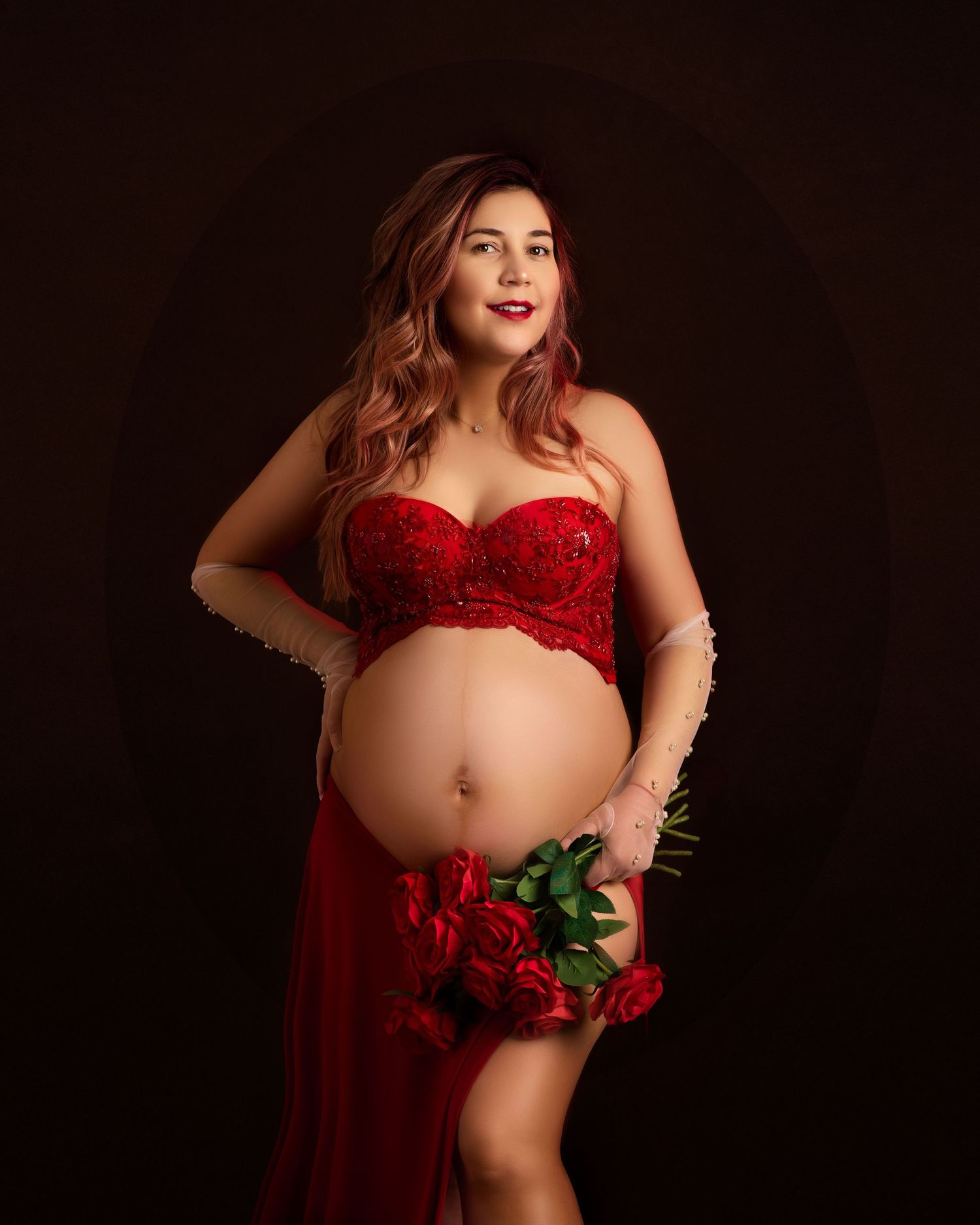 A pregnant woman in red lace lingerie holds red roses in a dark, romantic maternity photo.