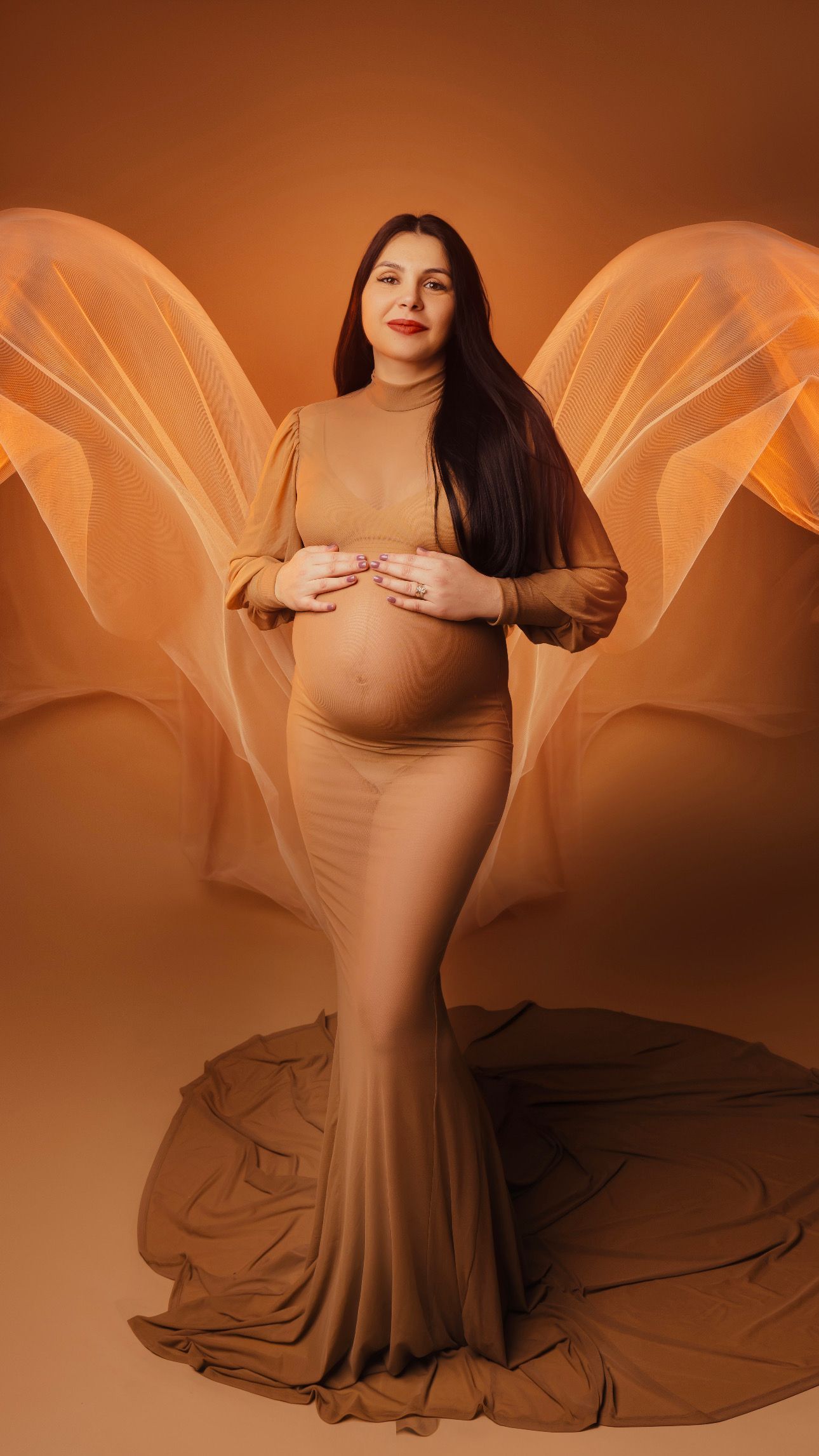 Maternity photo featuring an elegant form-fitting tan gown with ethereal wing-like background elements in warm amber tones.
