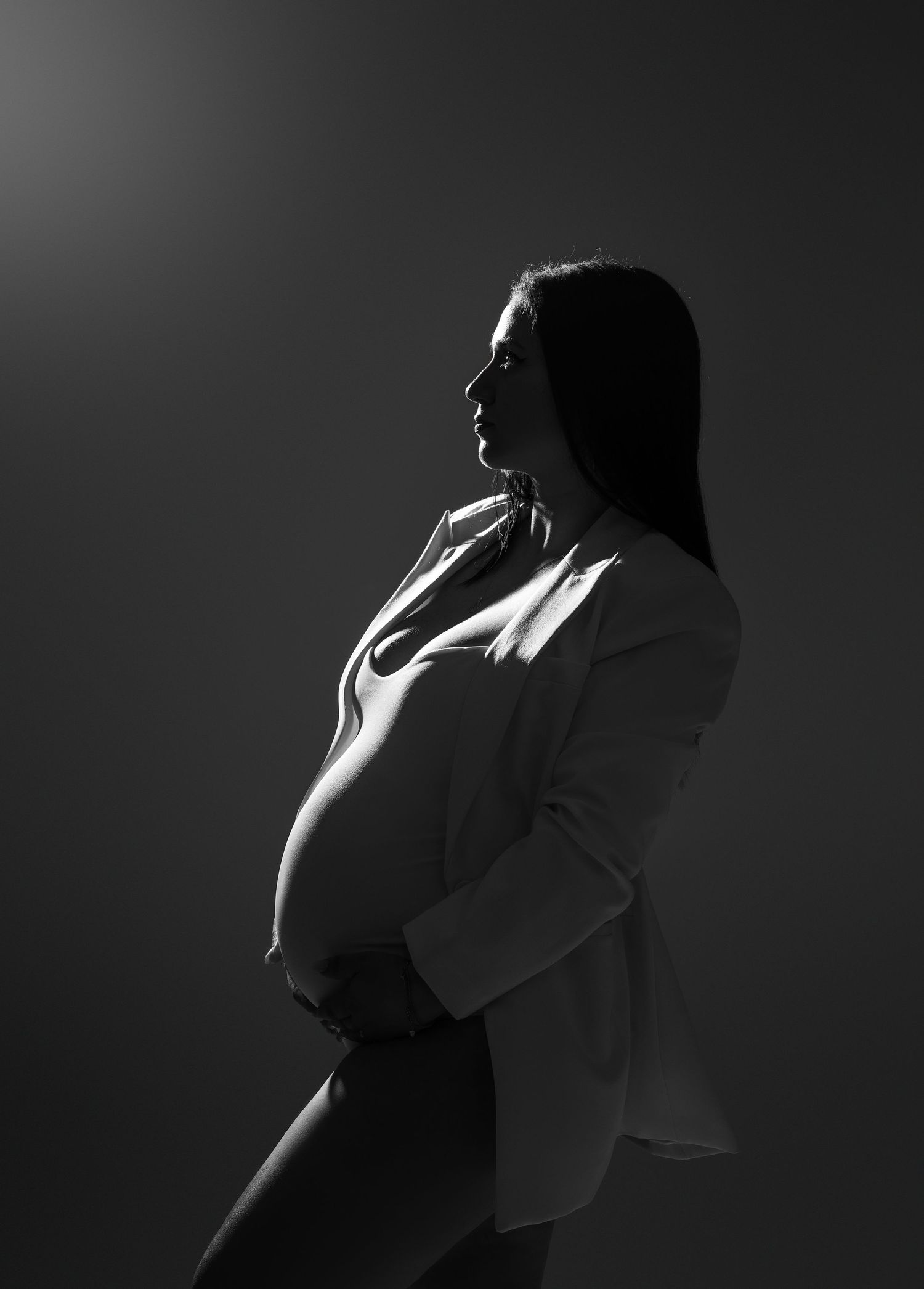 A dramatic black and white silhouette portrait of an expectant mother in profile against dark background.