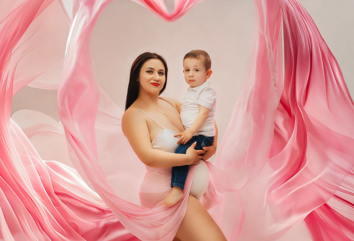 Flowing pink fabric creates a heart shape frame around parent and child in an elegant studio portrait.