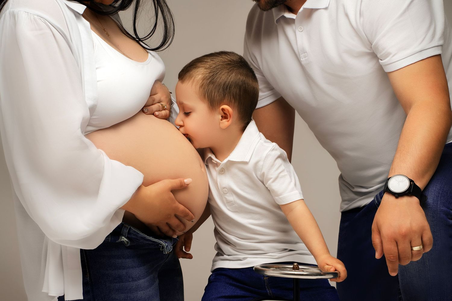 A young child in a white shirt kisses a pregnant belly while someone stands nearby wearing a wristwatch.