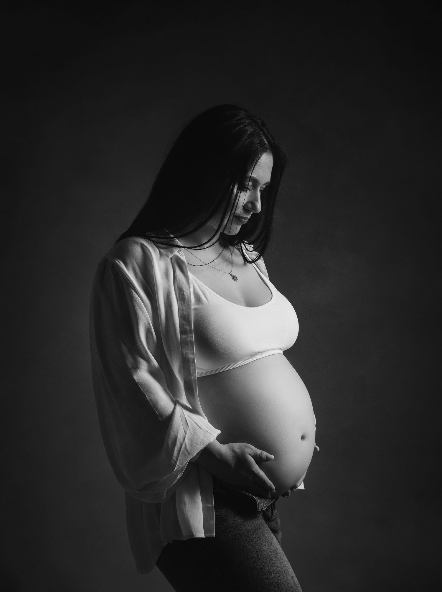 Black and white artistic maternity photograph with dramatic lighting showing an expectant mother in profile wearing white.