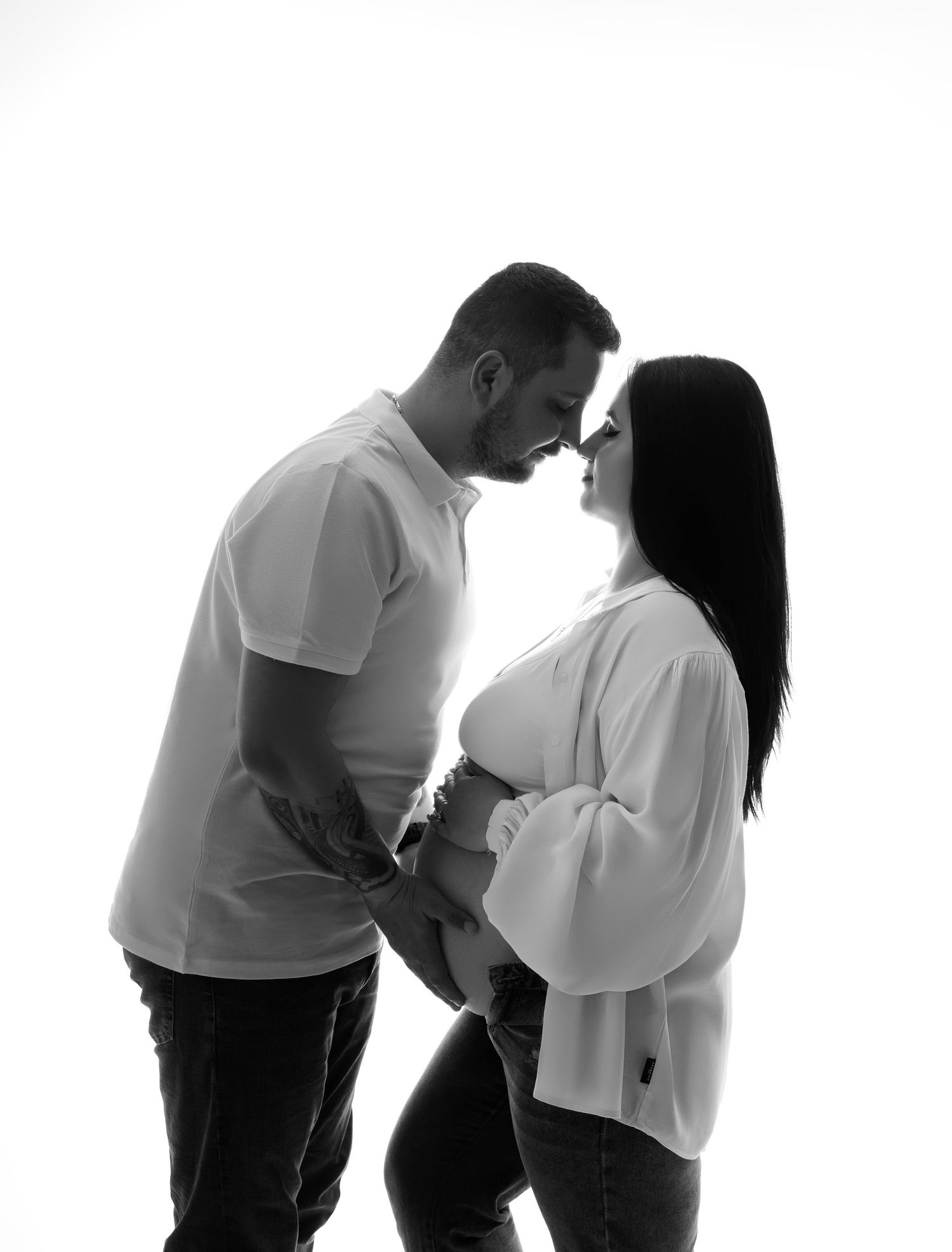 Black and white silhouette of an expecting couple in intimate pose wearing white tops against light background.
