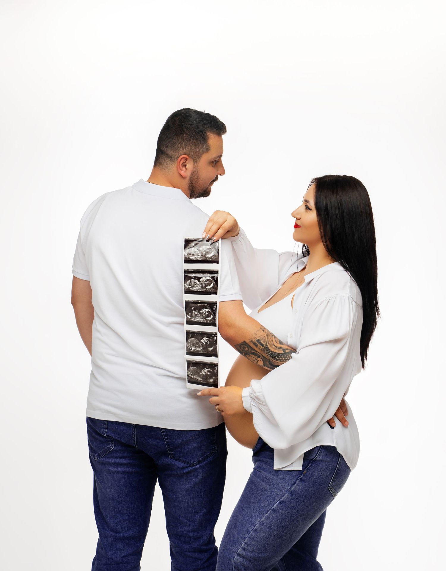 A couple in white tops and jeans hold up ultrasound photos while posing against a white background for a pregnancy announcement.