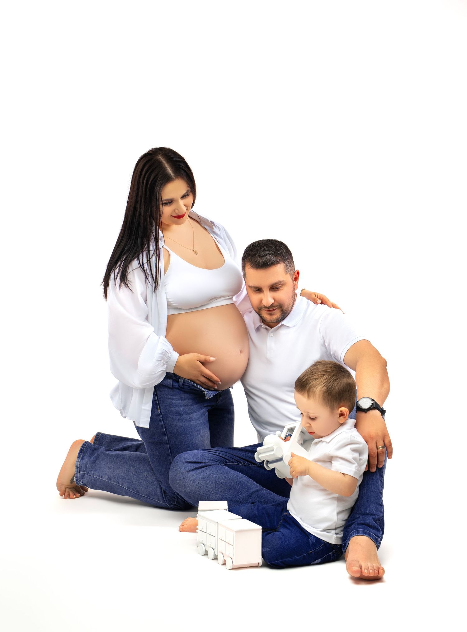 A pregnant woman in a white shirt and jeans sits with family on a white backdrop while looking at a young child playing.