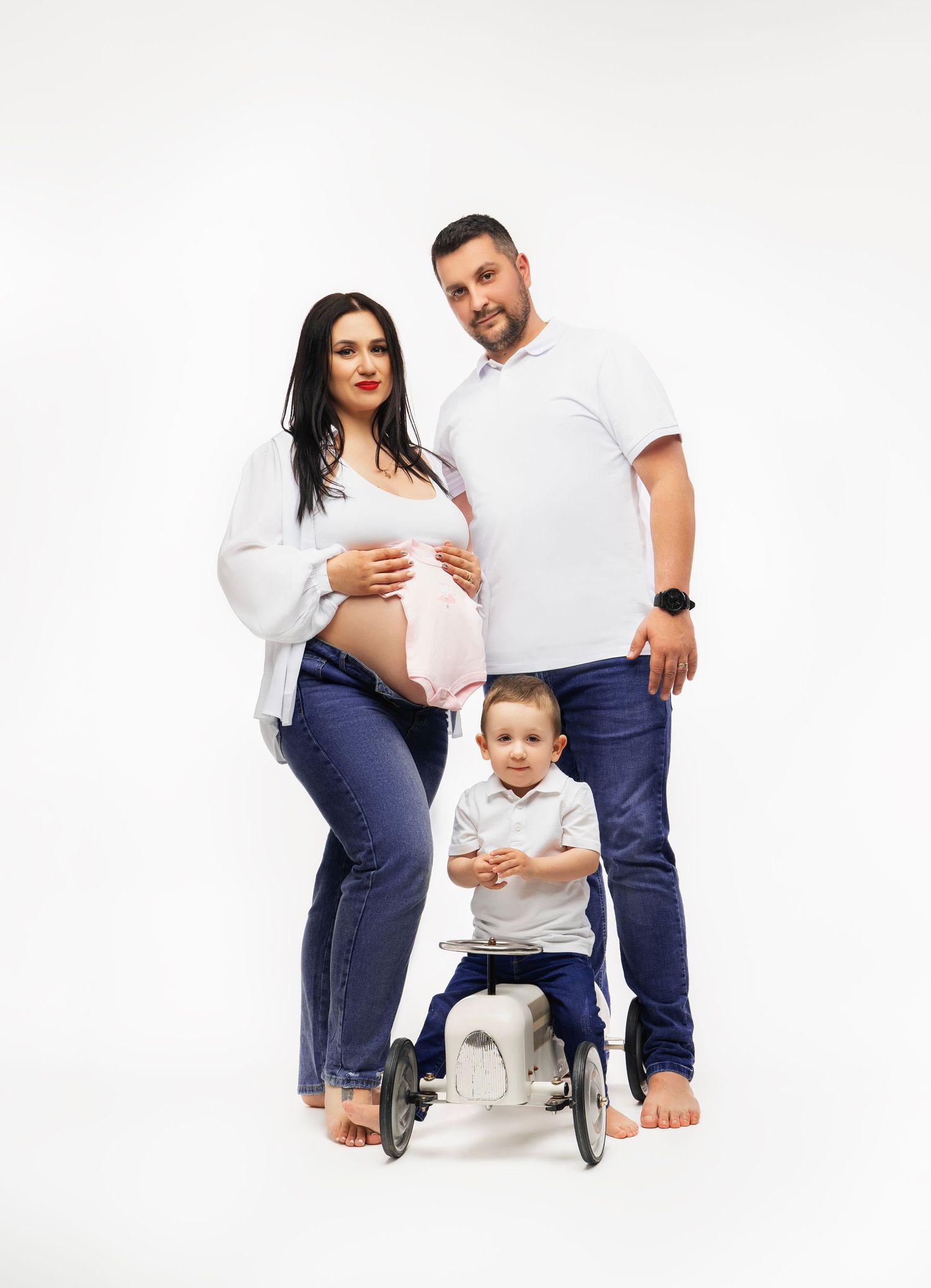Expectant family poses together in matching white and denim outfits against a white background for maternity photoshoot.