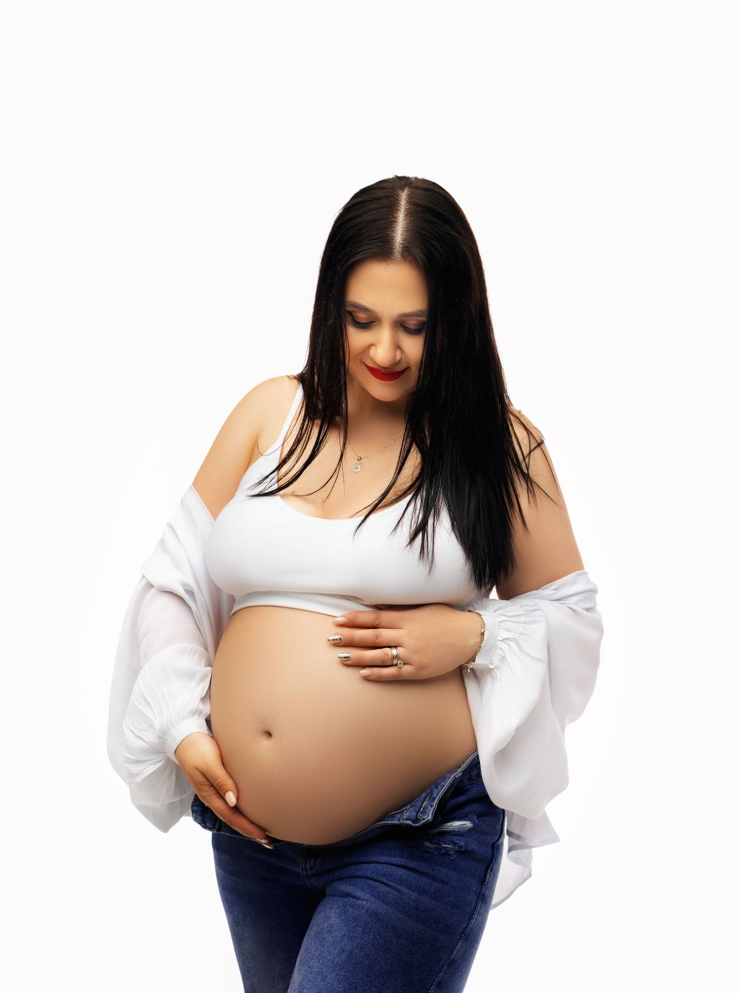 A pregnant woman wearing a white crop top and jeans gently touches her baby bump against a white background.